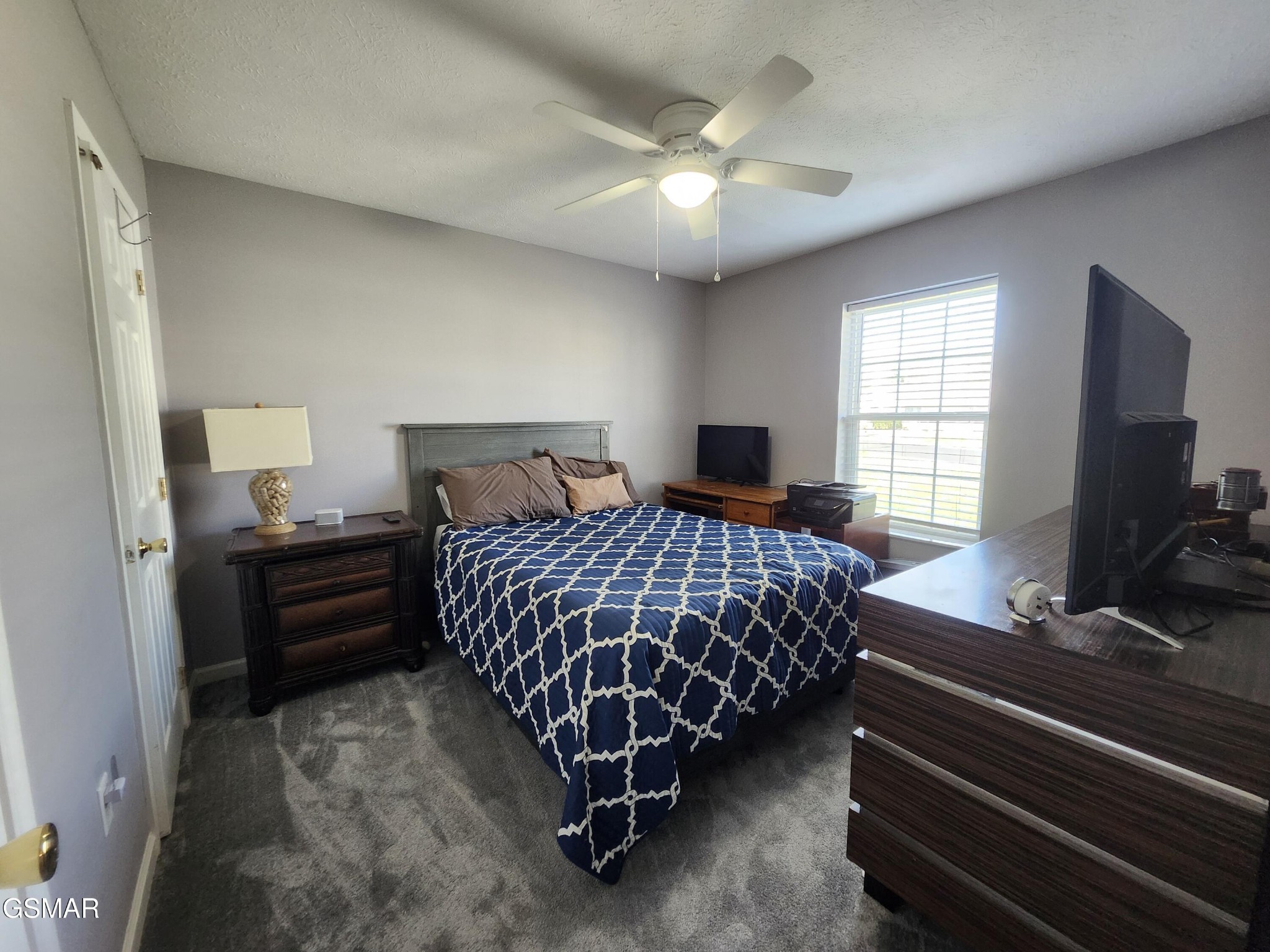 Furnished bedroom with a navy patterned bedspread, gray carpet, ceiling fan, and window with blinds.