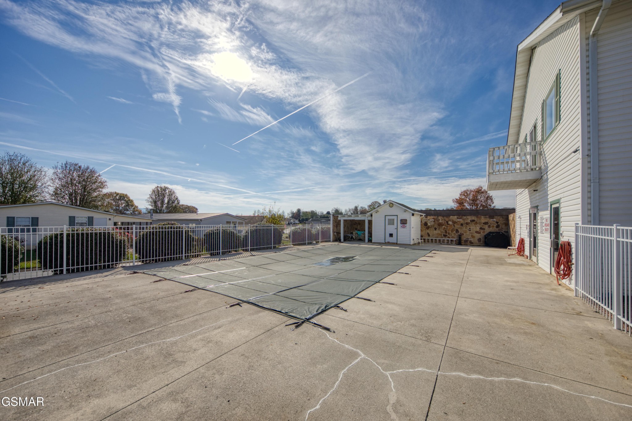 Outdoor swimming pool beside a white-sided building, with a covered pool and fenced concrete deck.