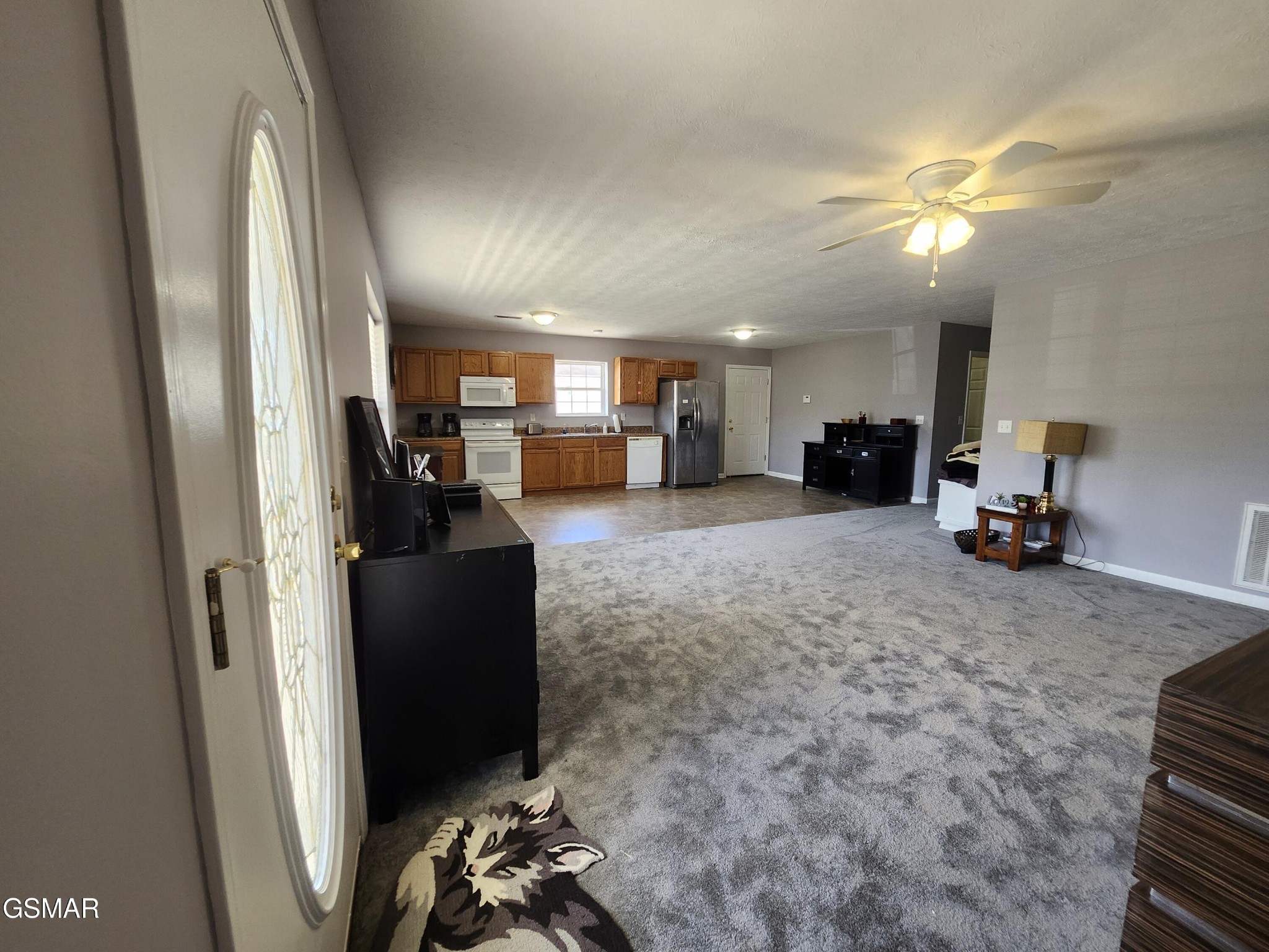 Open kitchen and living area with wood cabinets, stainless-steel refrigerator, and gray carpet.