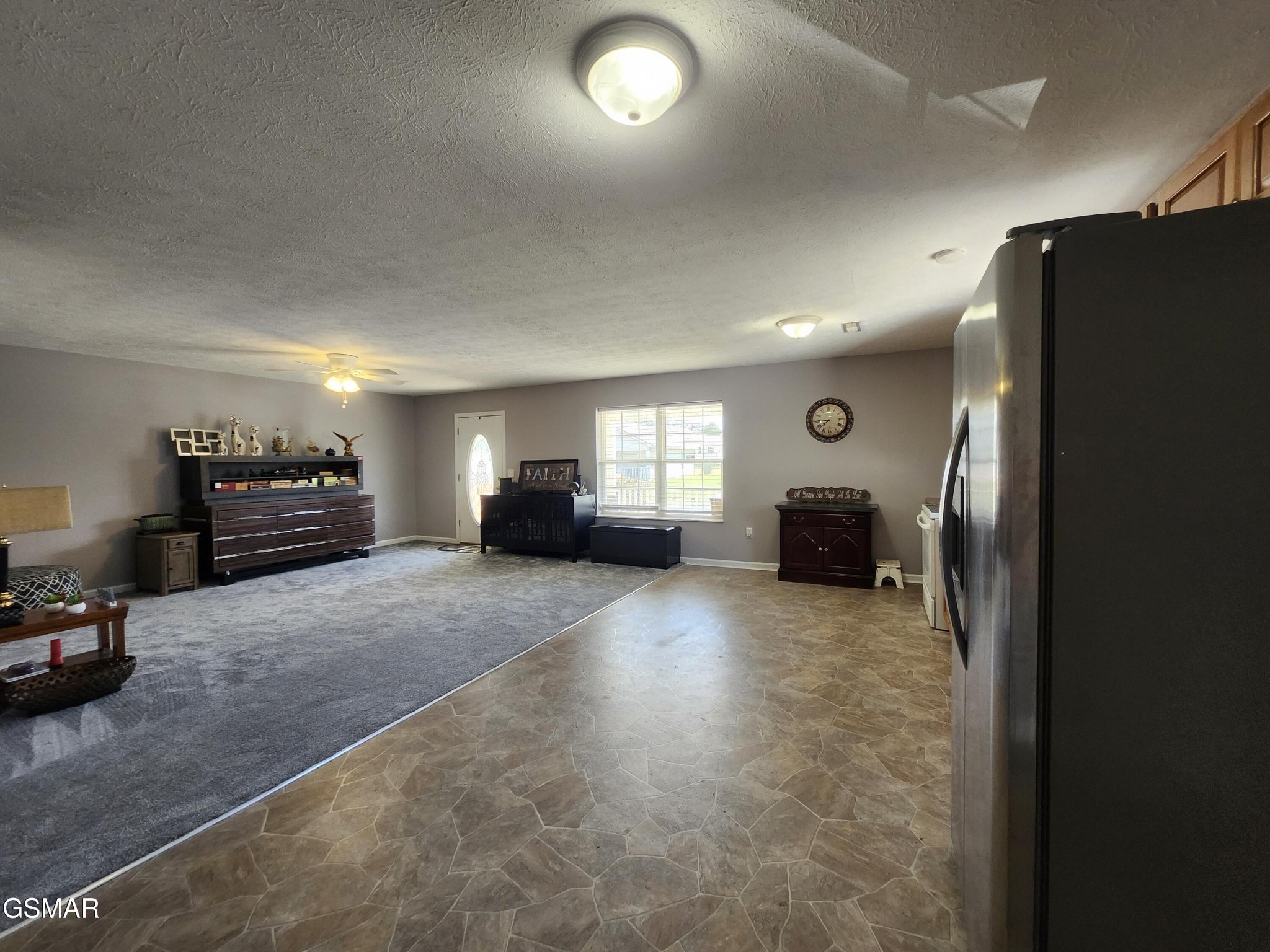 Furnished living area with gray carpet, tiled flooring, ceiling fan, and stainless-steel refrigerator.