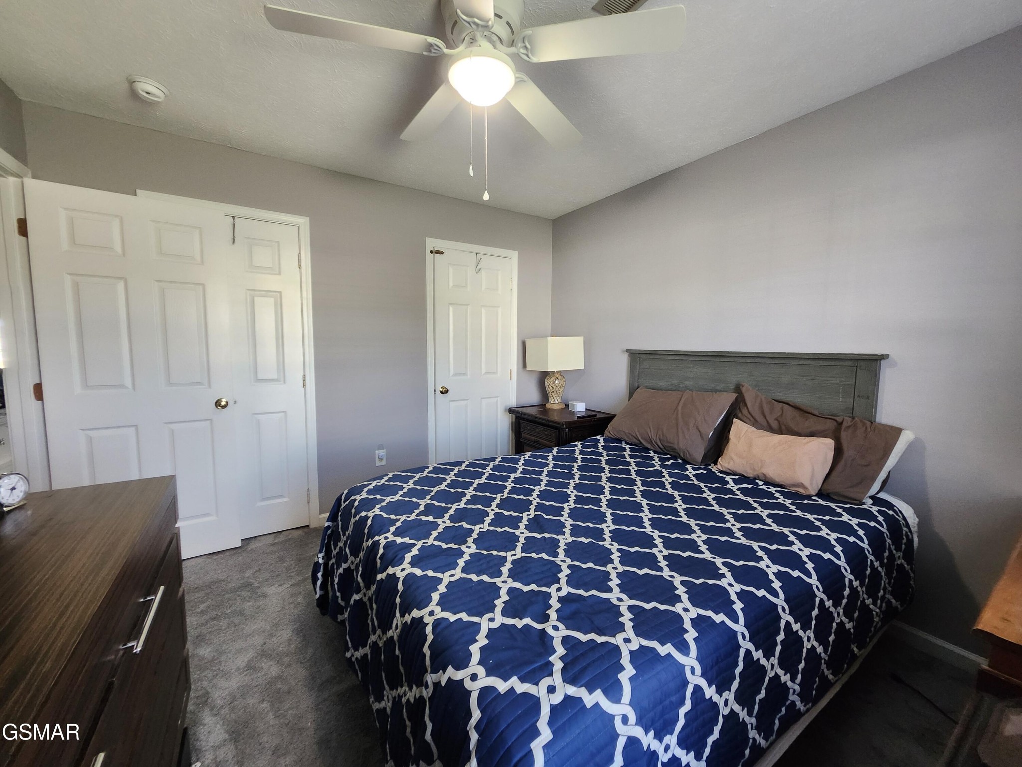 Furnished bedroom with a blue-and-white patterned bedspread, dark wood headboard, and ceiling fan.