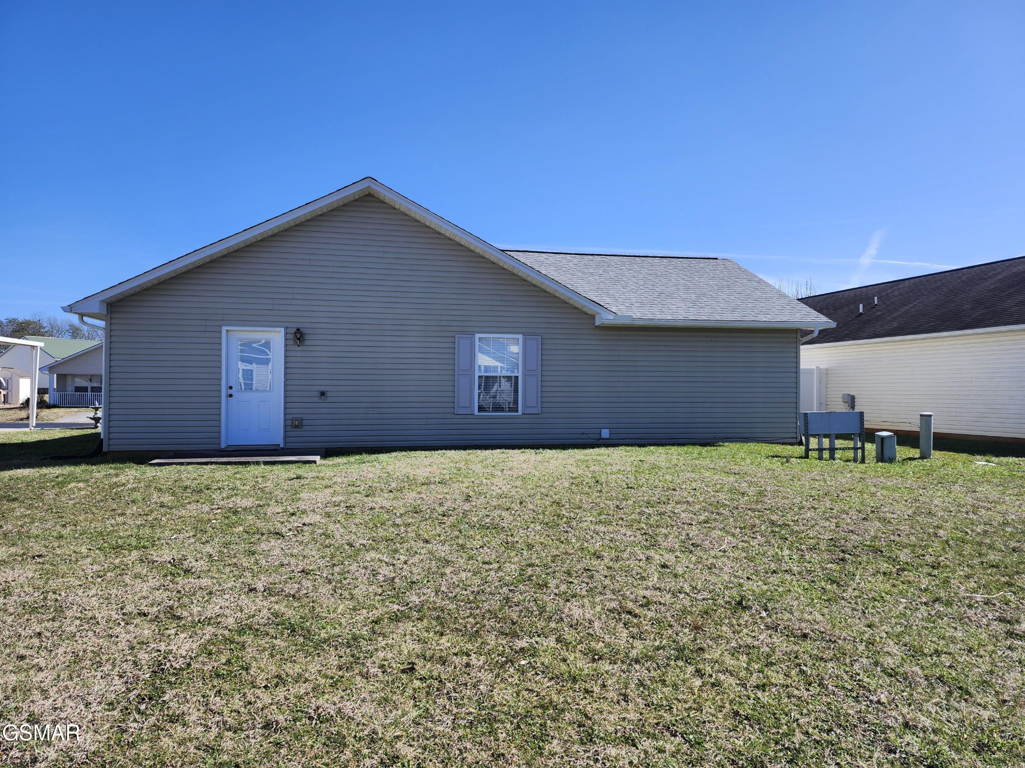 Gray single-story house rear exterior with a white door and shuttered window facing a broad grassy yard.