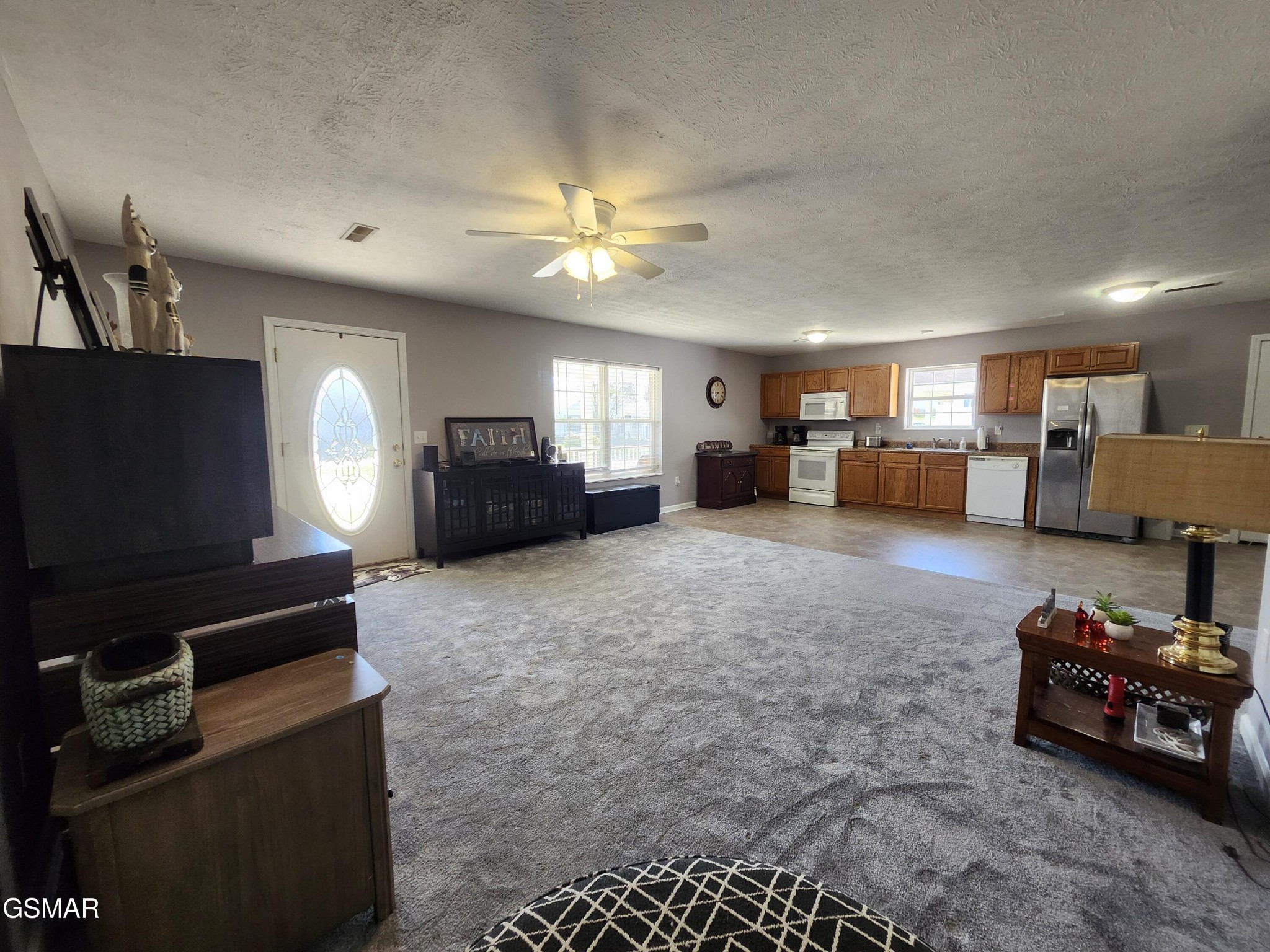 Open-plan living room and kitchen with gray carpet, wood cabinets, and stainless steel appliances.
