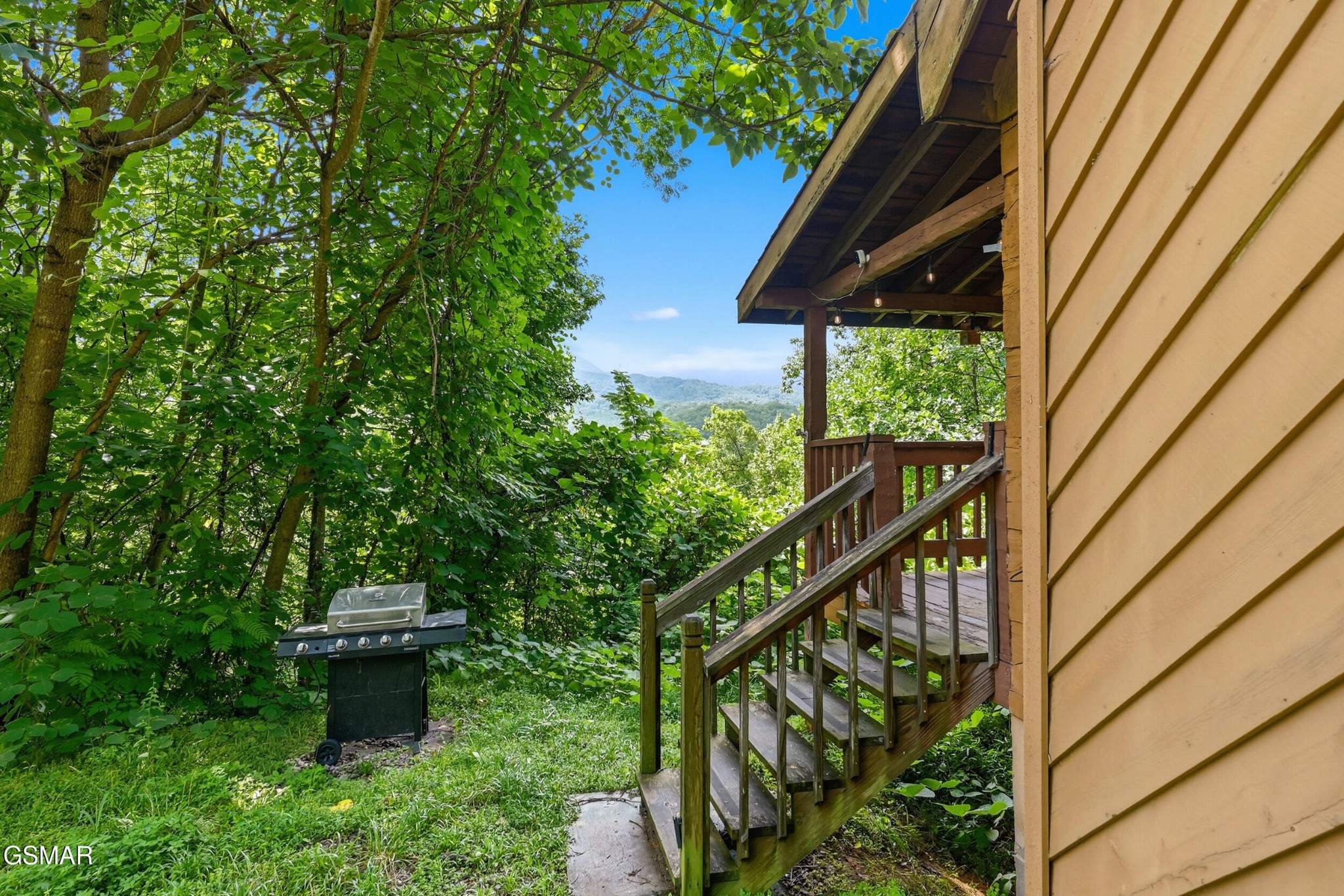Cabin porch and stairs beside a grill, surrounded by trees with a mountain view.