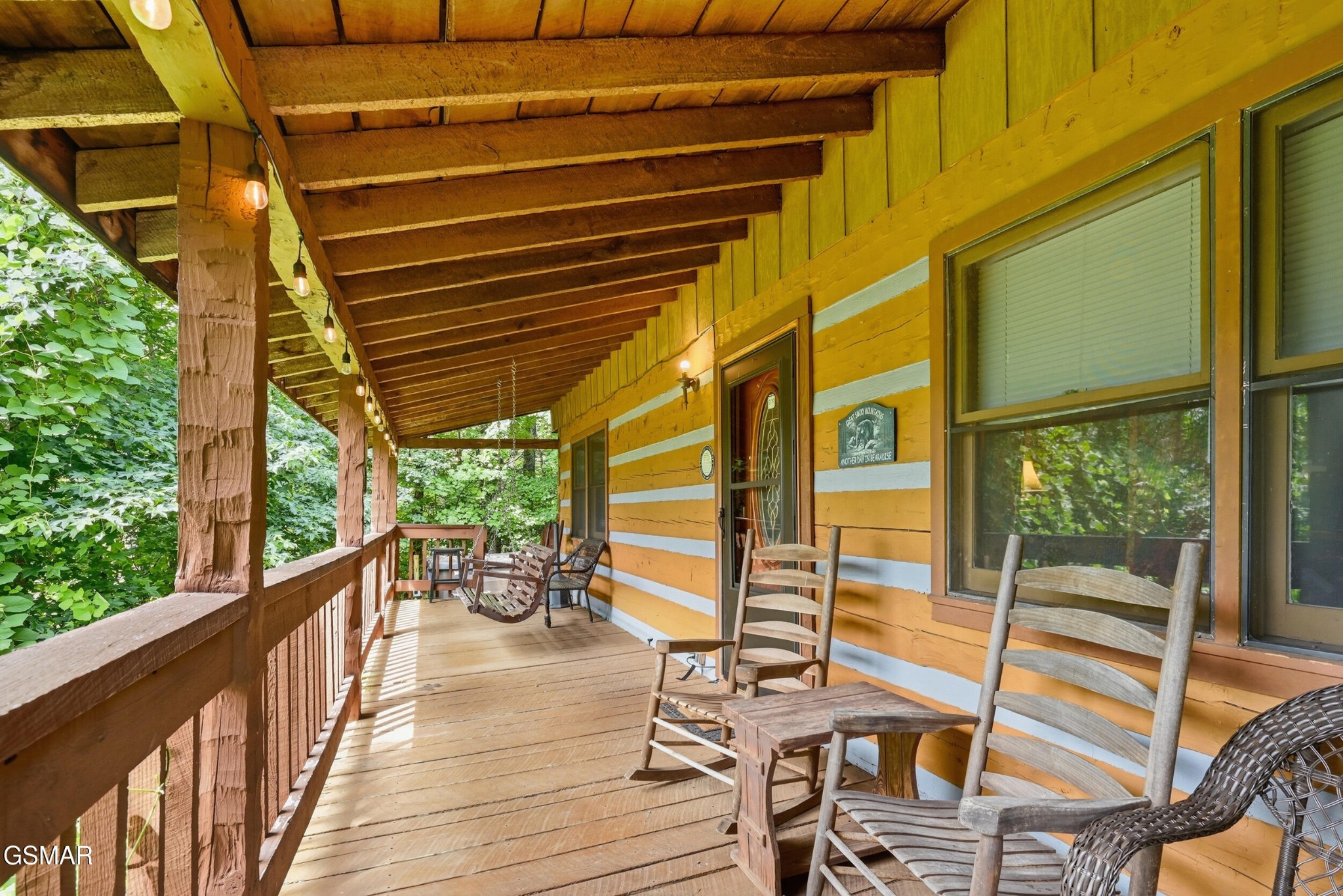 Covered wooden porch with rocking chairs, string lights, and wooded surroundings.