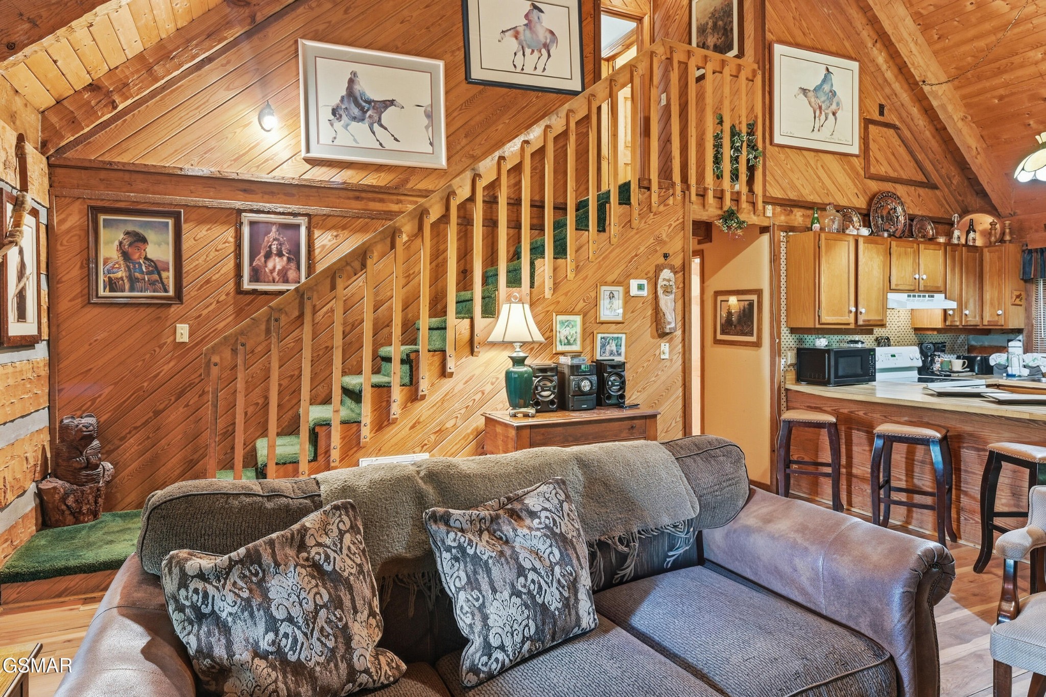 Cabin living room with a wood-paneled vaulted ceiling, green-carpeted staircase, and open kitchen.