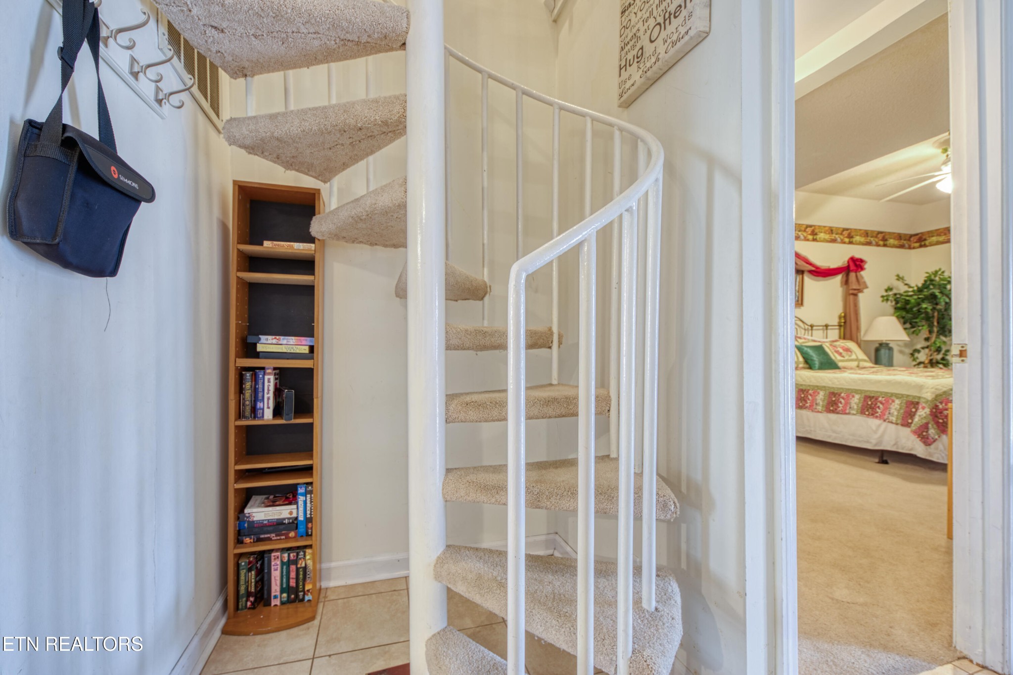 Carpeted spiral staircase with white railing beside a bookcase and open bedroom doorway.