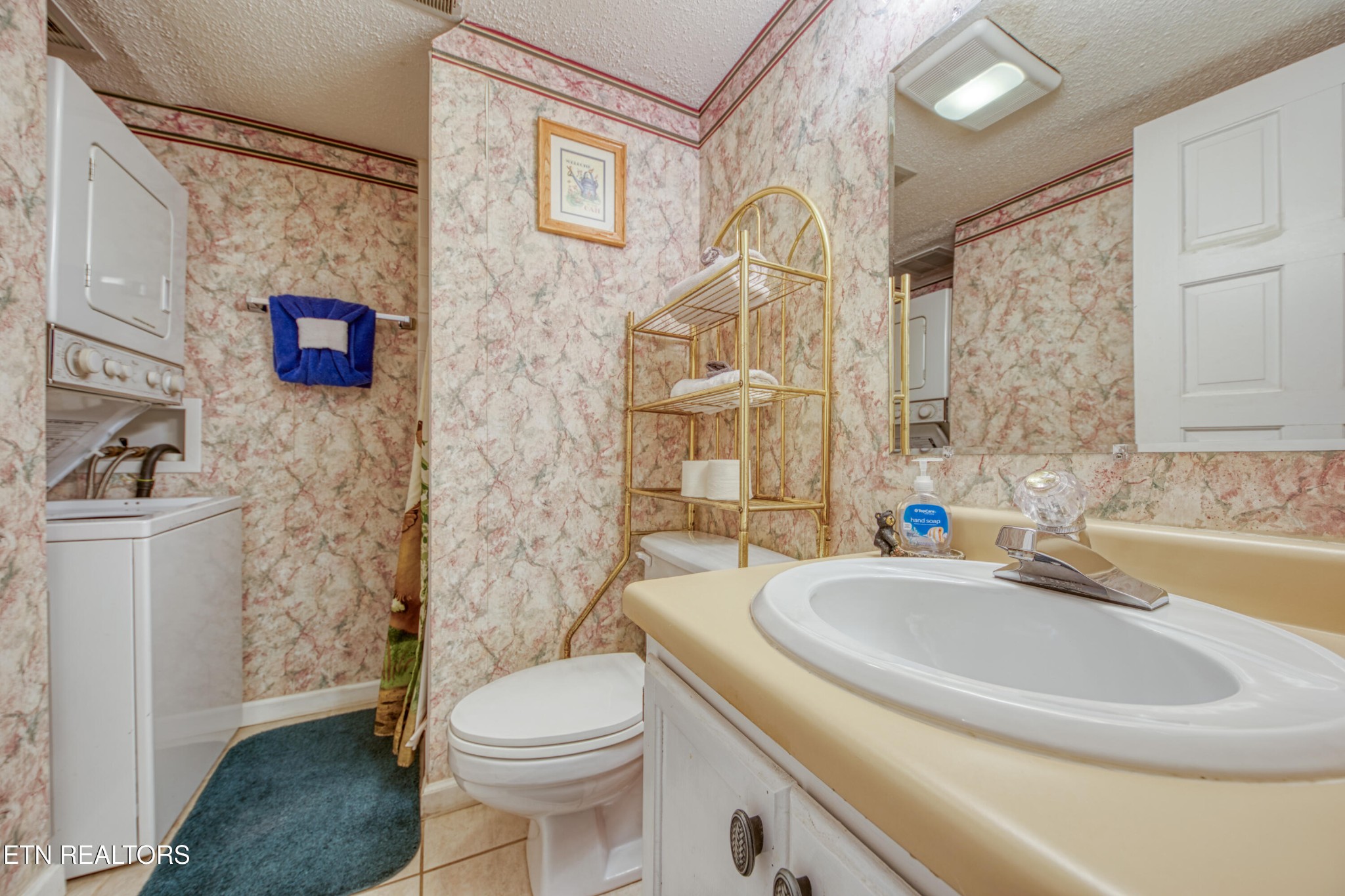 Bathroom with pink marbled wallpaper, a white vanity, toilet, and stacked washer and dryer.
