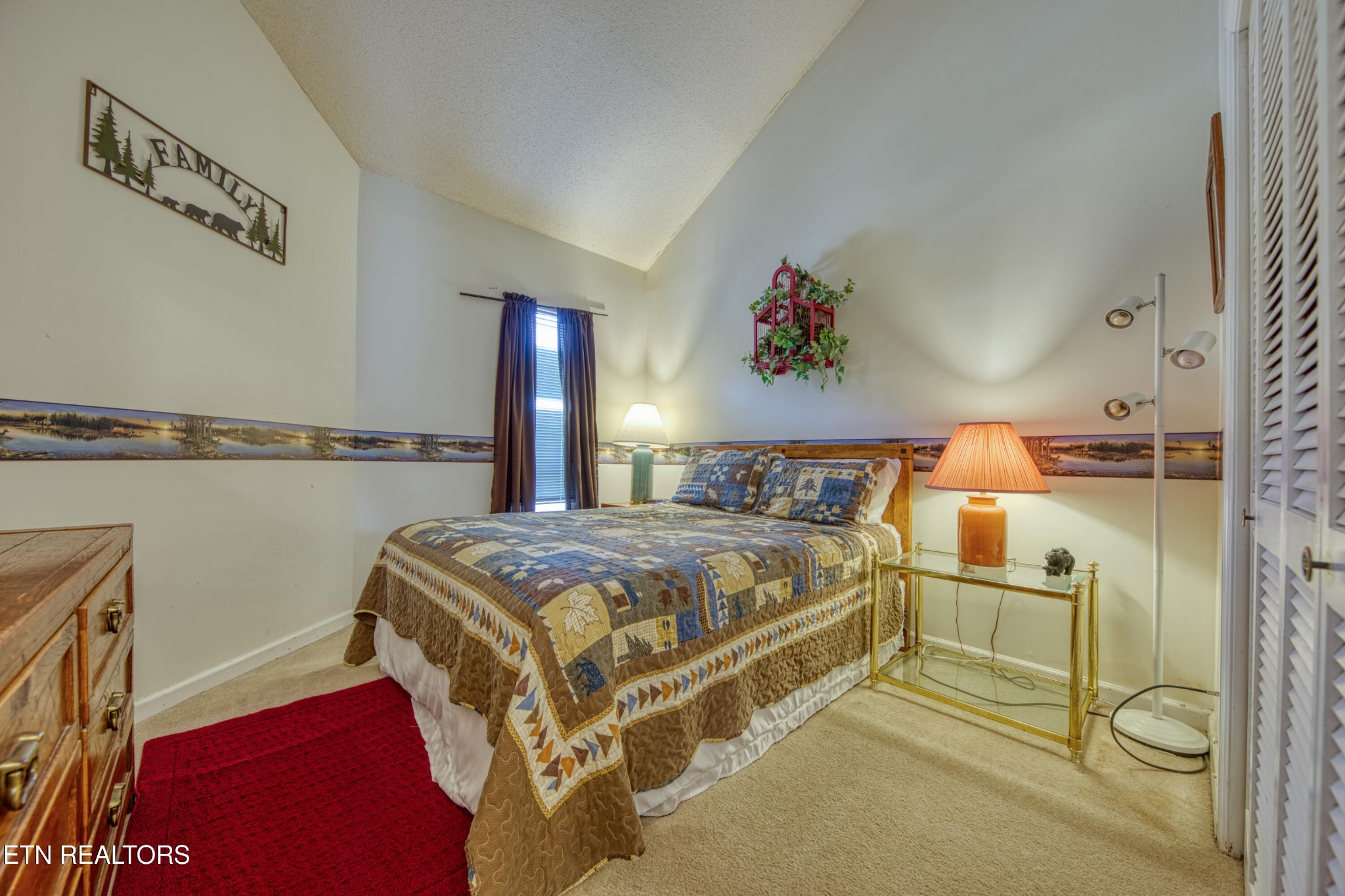 Furnished bedroom with a patchwork quilt, red rug, and louvered closet doors.