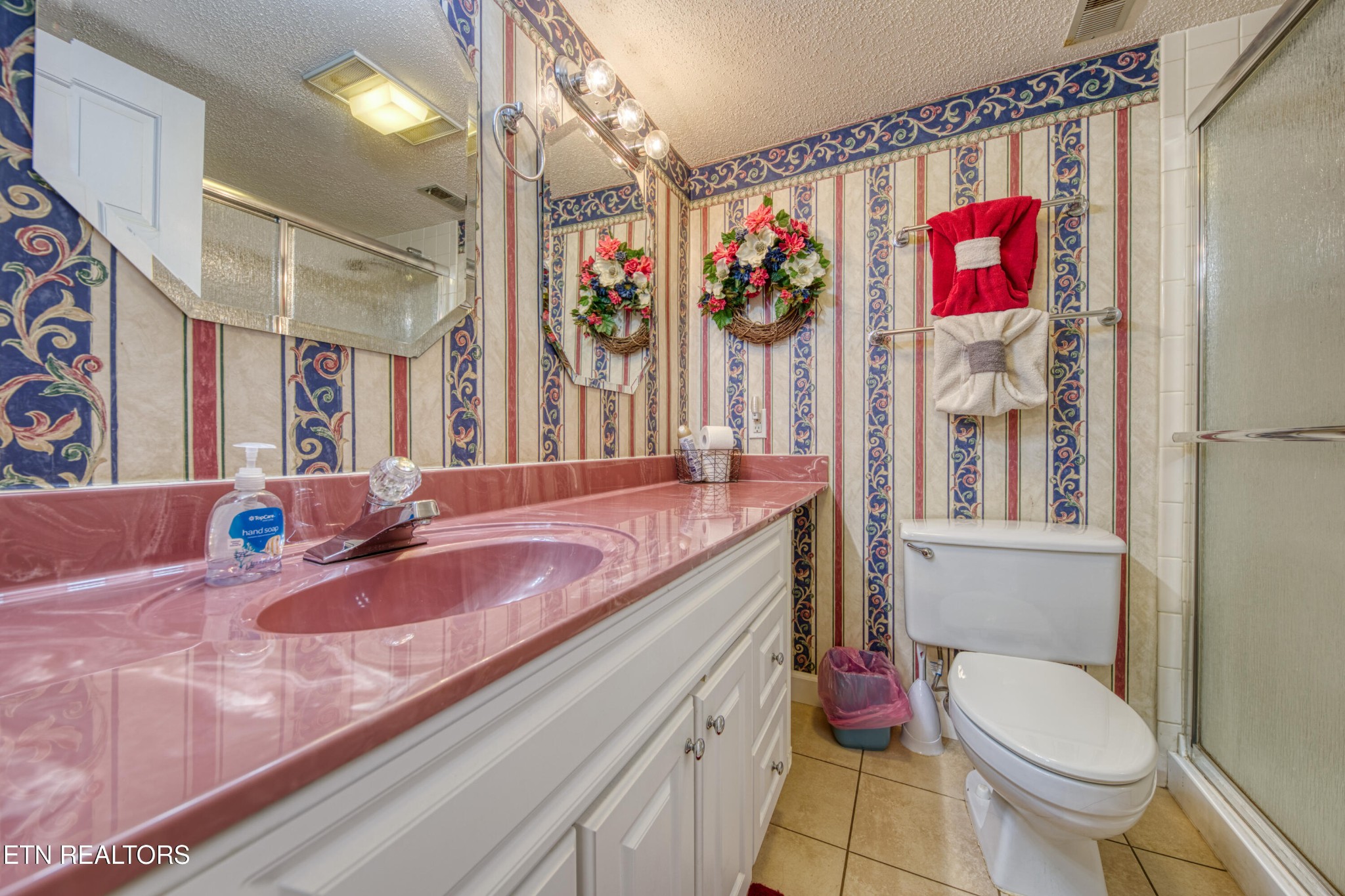 Bathroom with a pink sink counter, ornate striped wallpaper, white vanity, toilet, and glass shower.