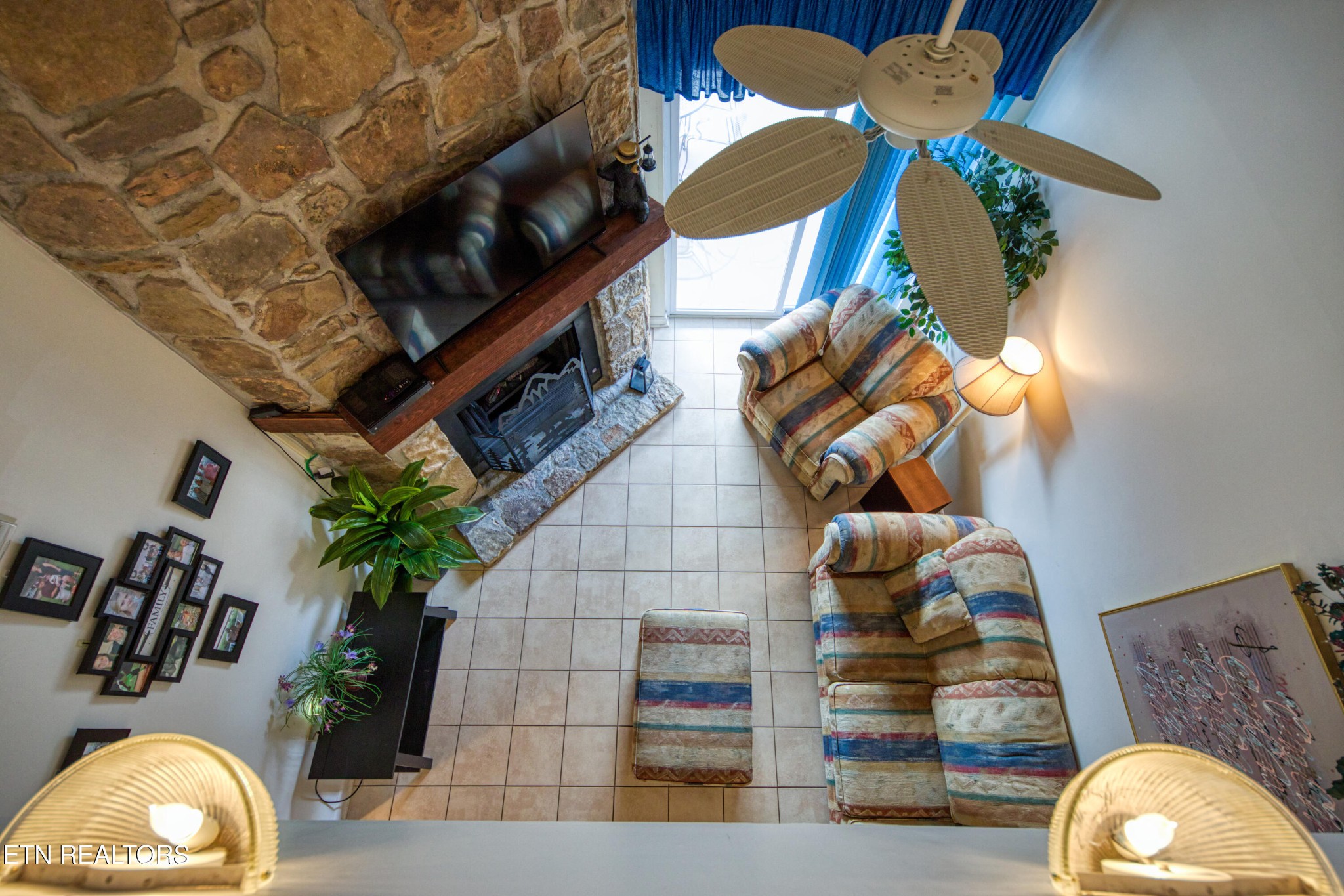 Furnished living room with a stone fireplace wall, striped seating, and a ceiling fan.