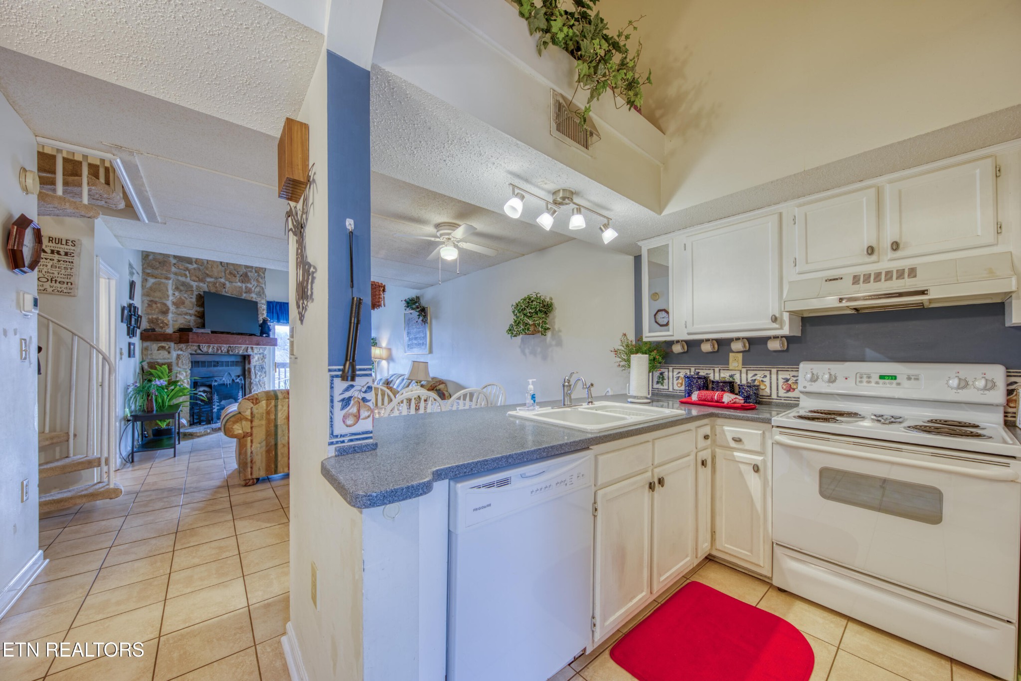 Kitchen with white cabinets and gray counters, opening onto a living room with a stone fireplace.