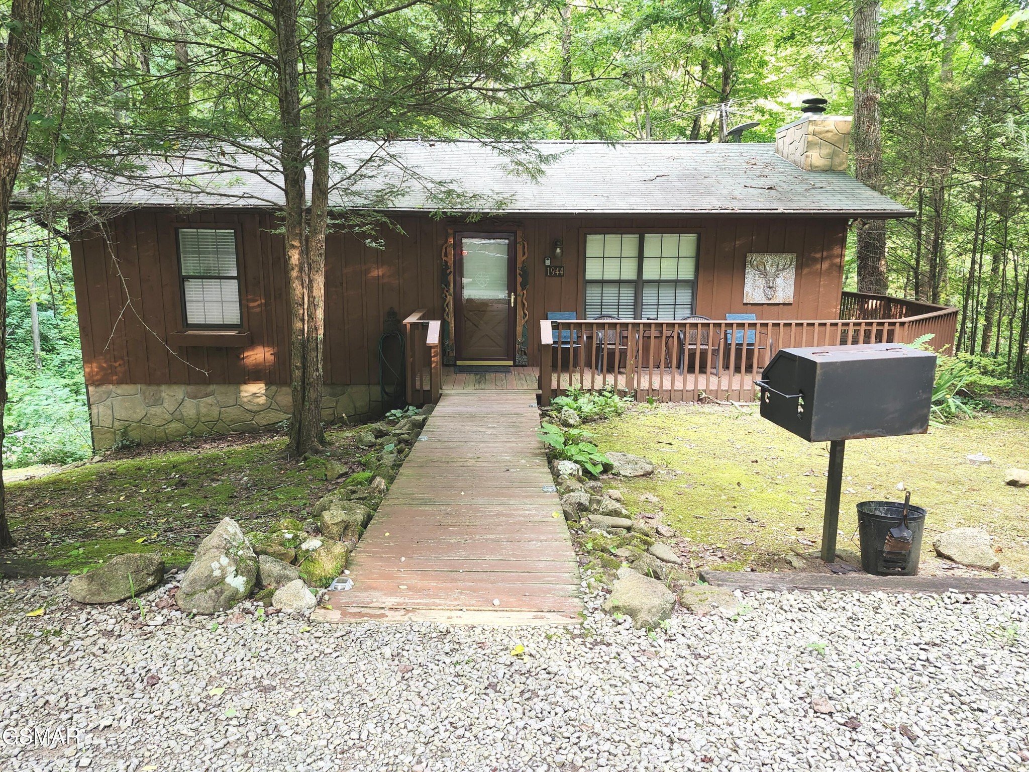 Woodland cabin with a stone foundation, covered deck, and wooden walkway across a gravel yard.