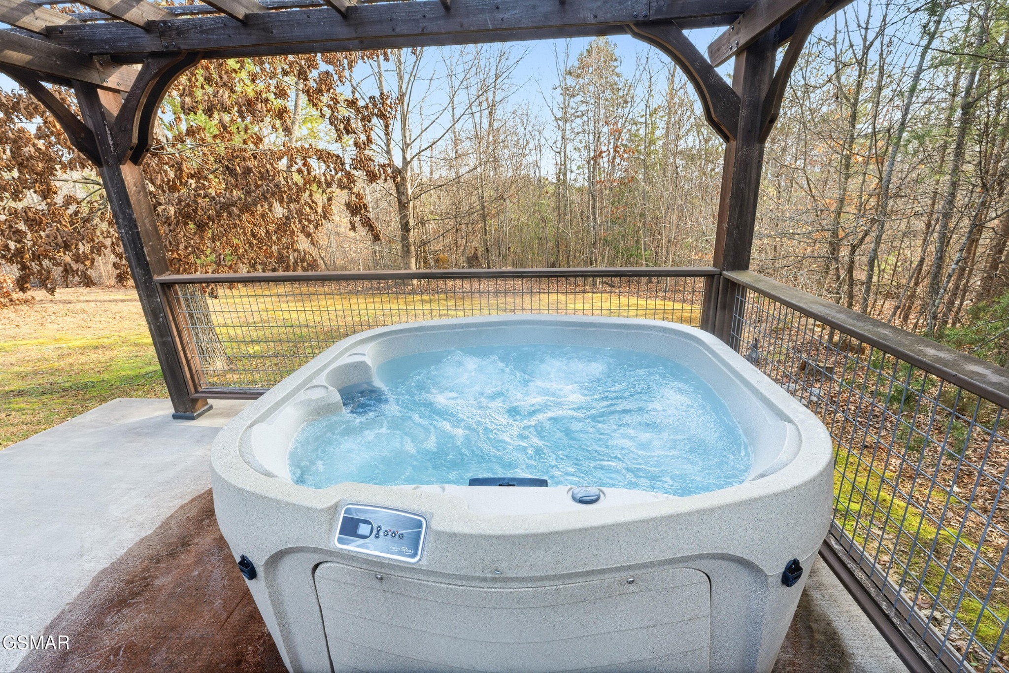 Outdoor hot tub beneath a wooden pergola, overlooking a wooded yard.