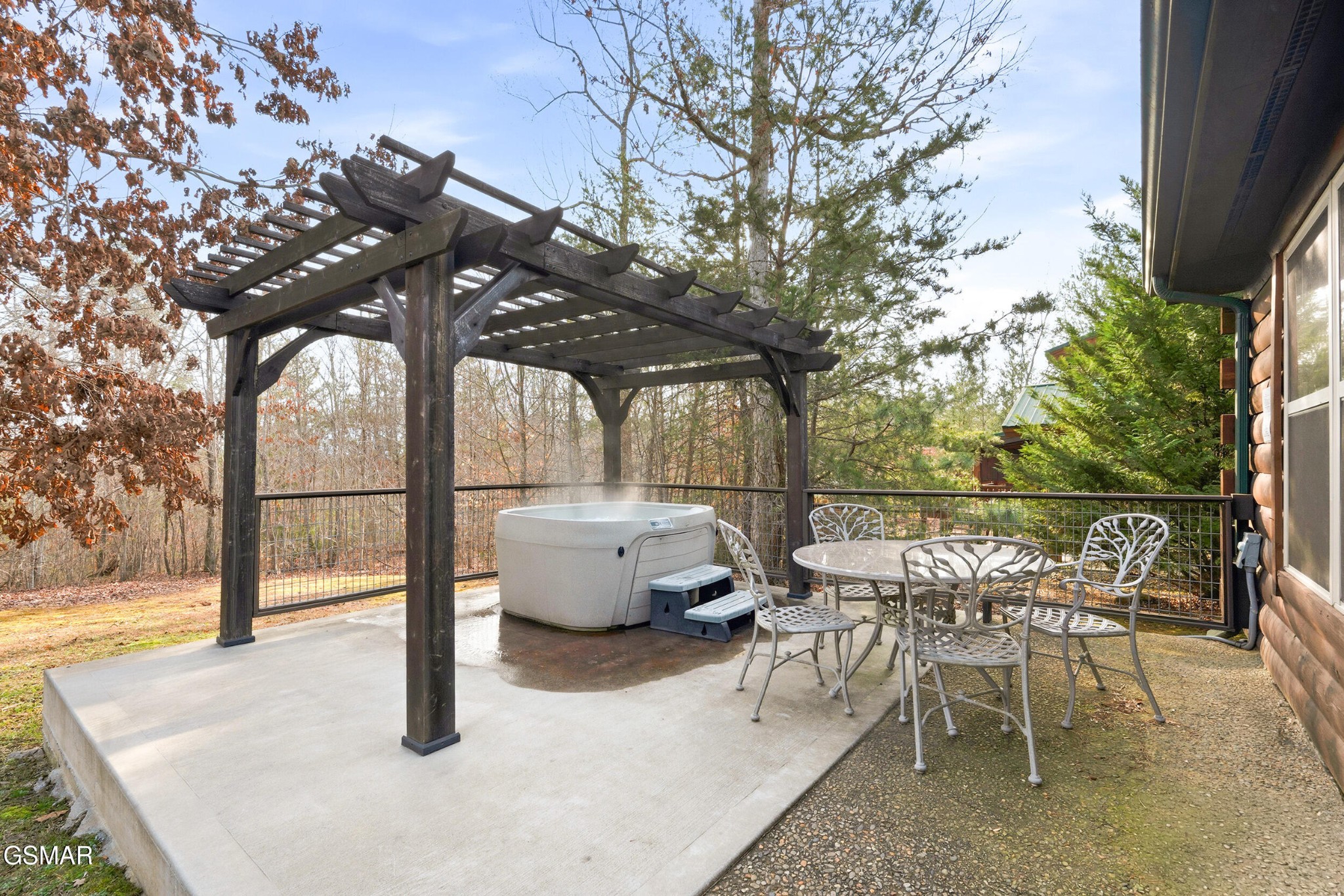 Patio with a hot tub beneath a wooden pergola, dining set, and wooded surroundings.