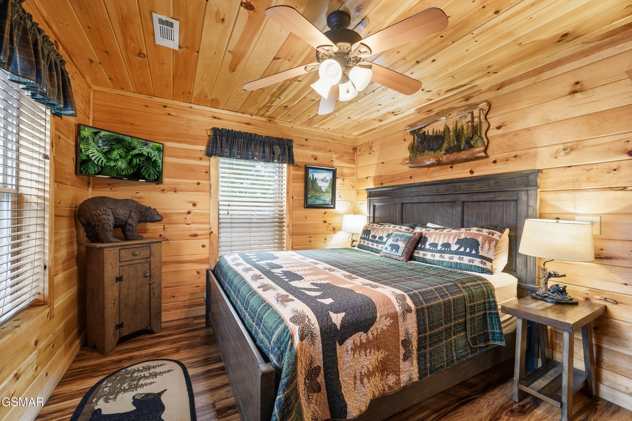 Furnished cabin bedroom with wood-paneled walls, a patterned bedspread, and bear-themed decor.