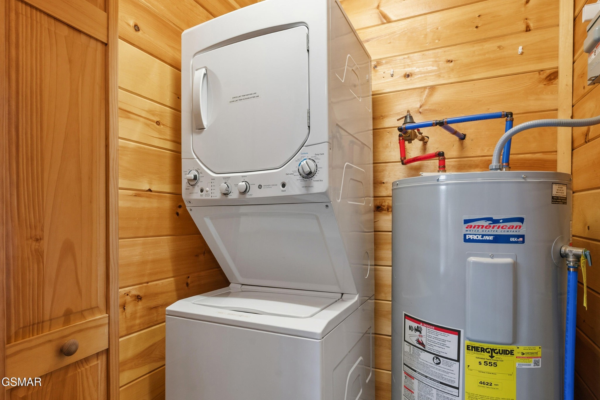 Laundry room with a white stacked washer-dryer, gray water heater, and wood-paneled walls.