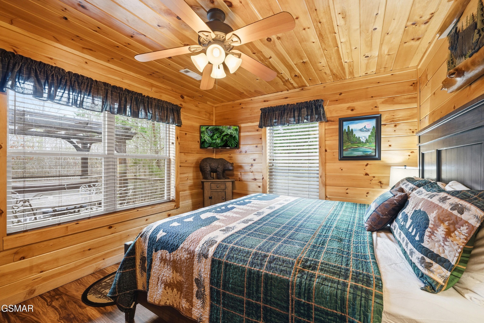 Wood-paneled bedroom with a blue quilted bed, ceiling fan, and windows overlooking a deck.