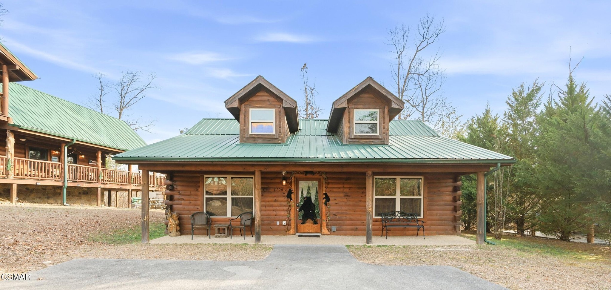 Log cabin with a green metal roof, two dormers, and a covered porch beside a second log building.