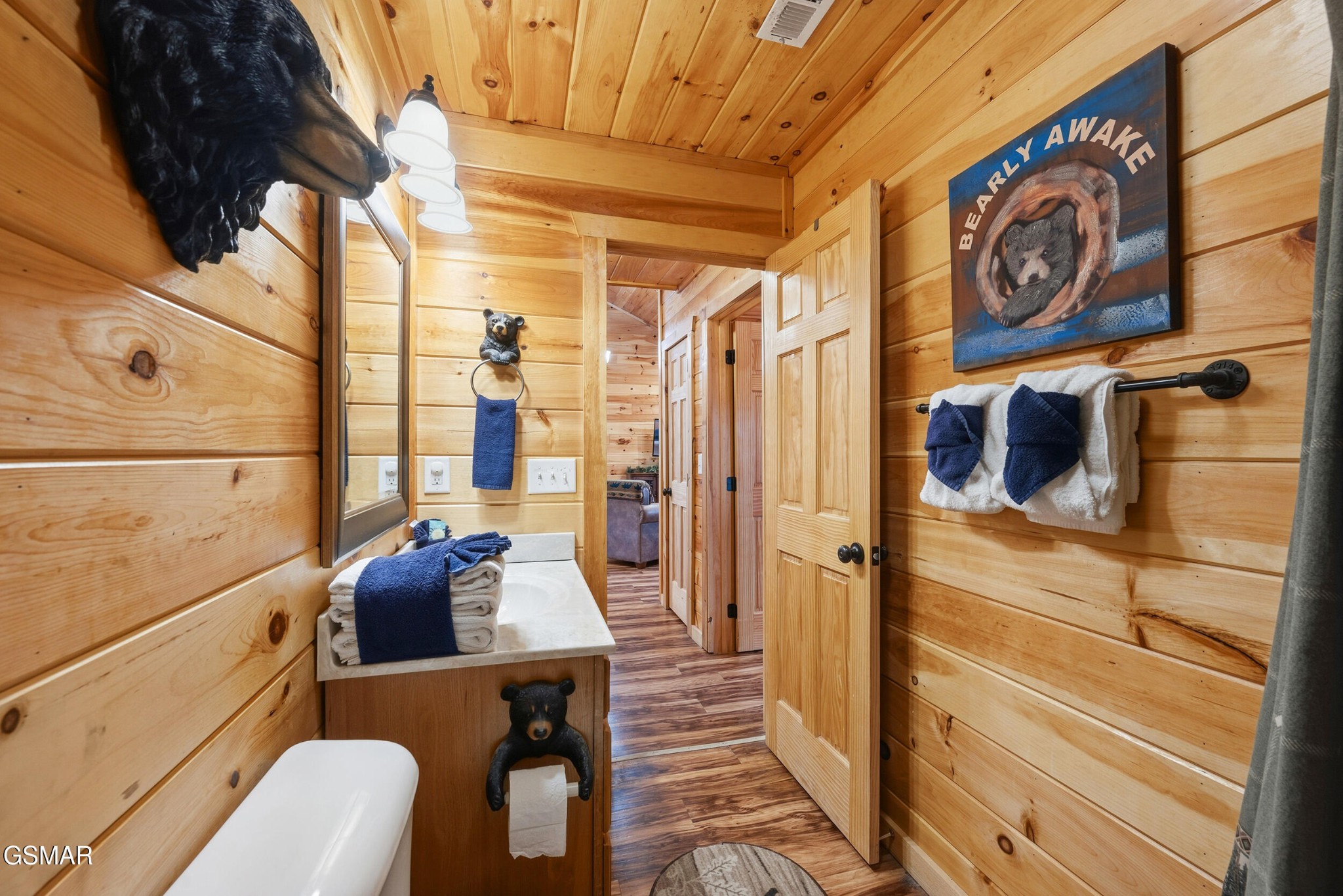 Rustic pine-paneled bathroom with a wood vanity, blue towels, bear decor, and an open doorway.