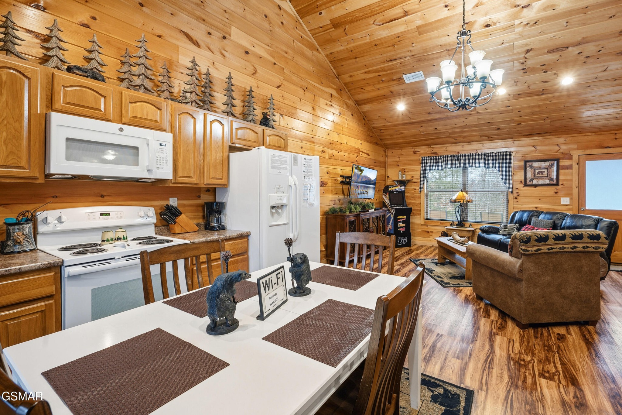 Open-plan cabin kitchen and living room with vaulted wood ceilings, white appliances, and bear-themed decor.