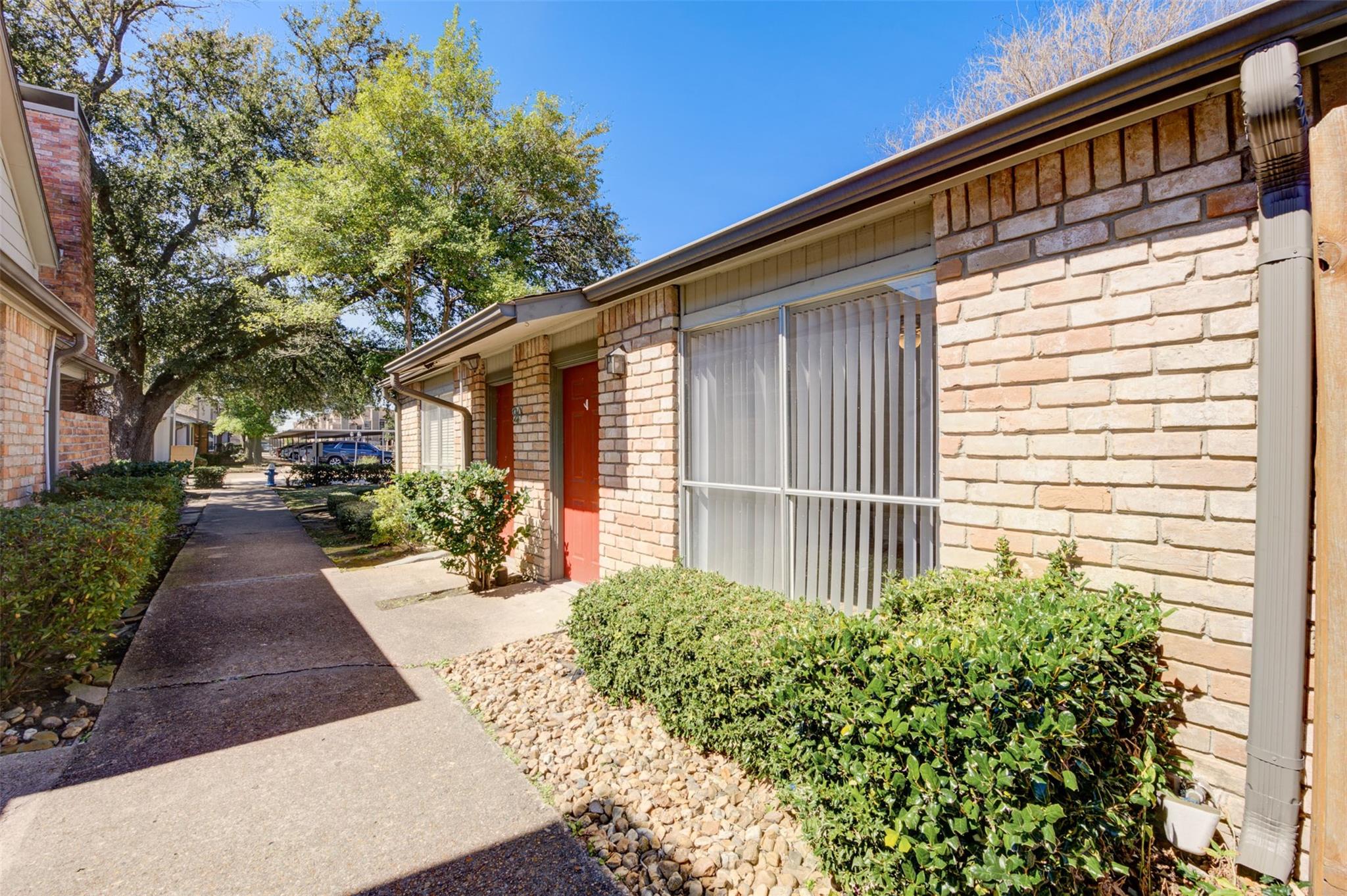Brick condominium exterior with red entry doors, barred windows, shrubs, and a shared walkway.
