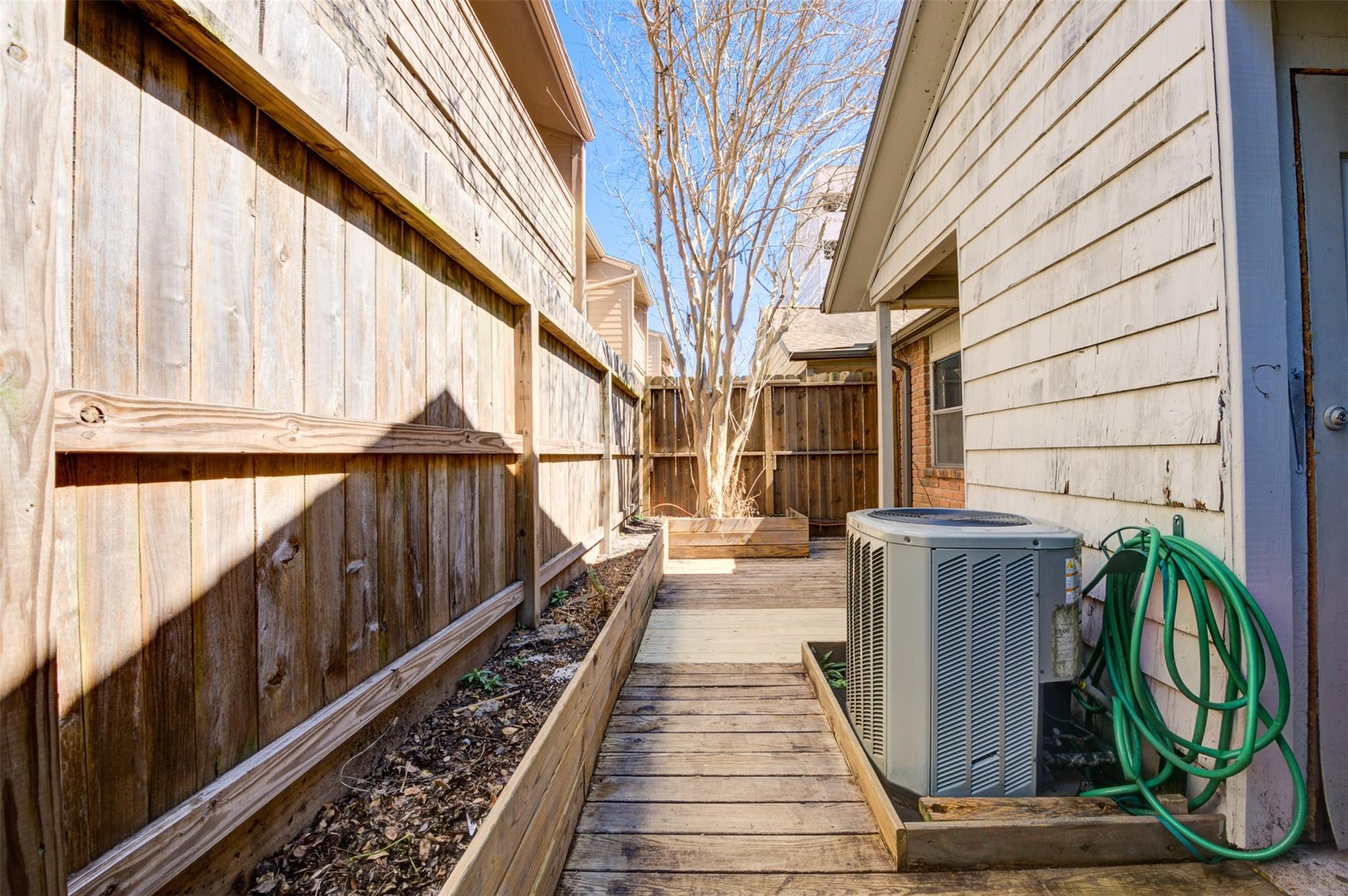 Townhouse side yard with a wooden walkway, raised beds, privacy fence, and coiled green hose.