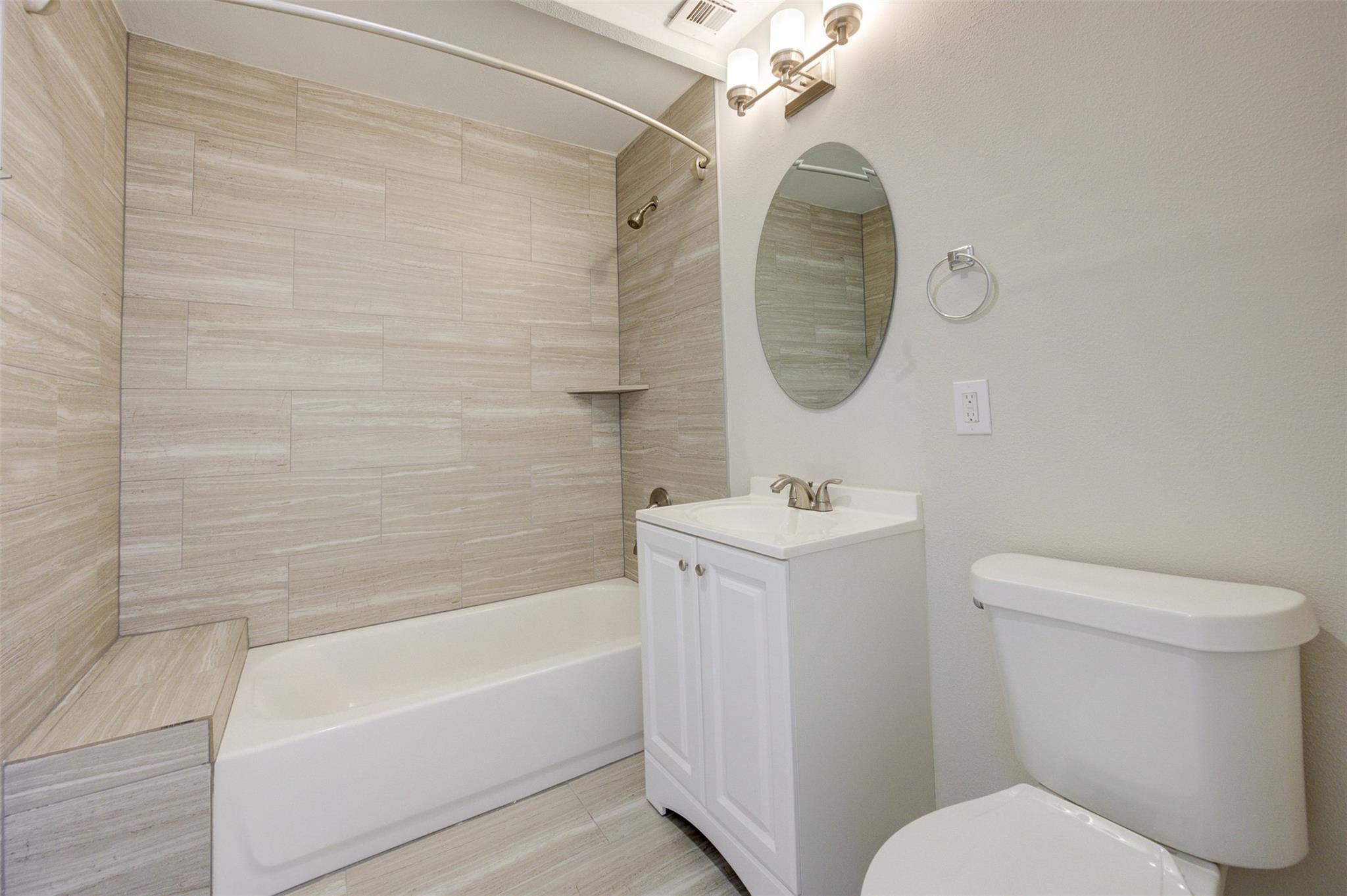 Bathroom with a large beige tile tub surround, white vanity, oval mirror, and toilet.