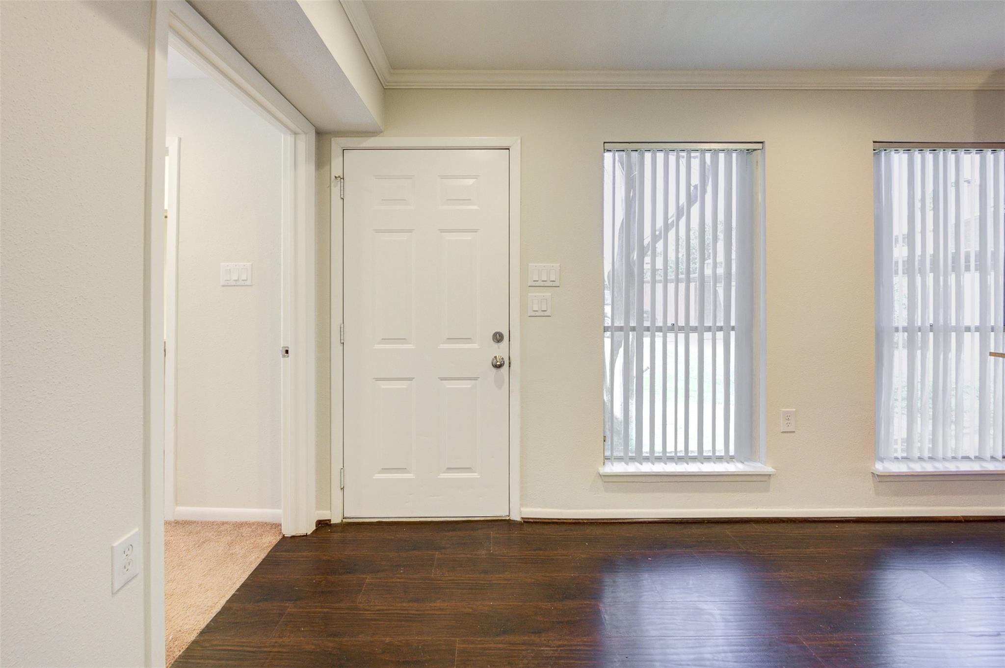 Unfurnished condo entry with a white paneled door, dark wood floors, and two windows with vertical blinds.