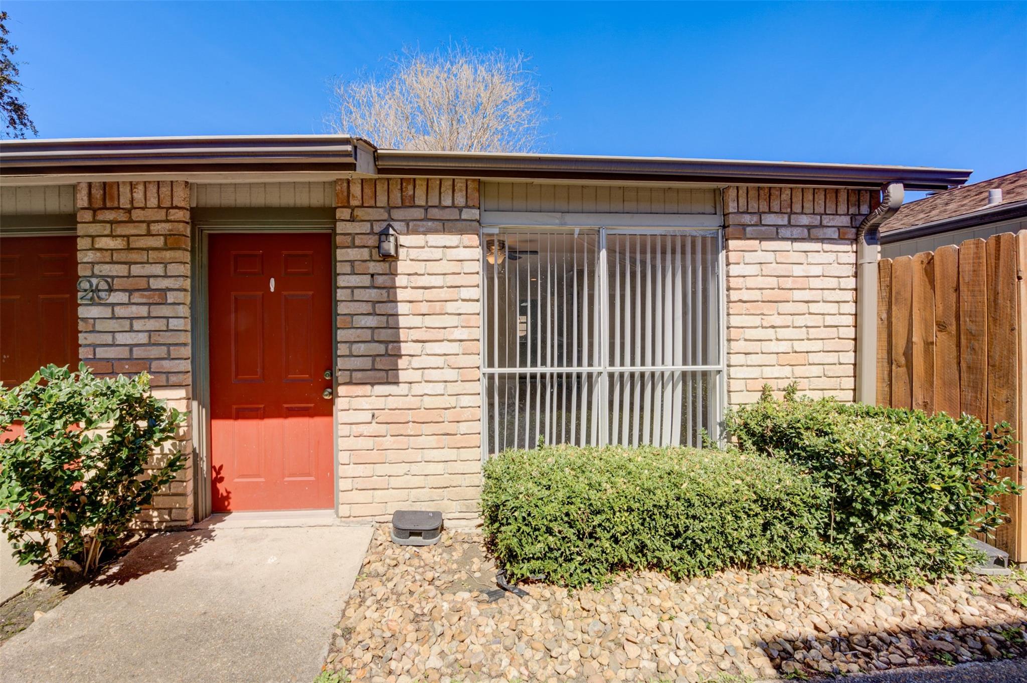 Brick home exterior with a red front door, barred window, trimmed shrubs, and wooden fence.