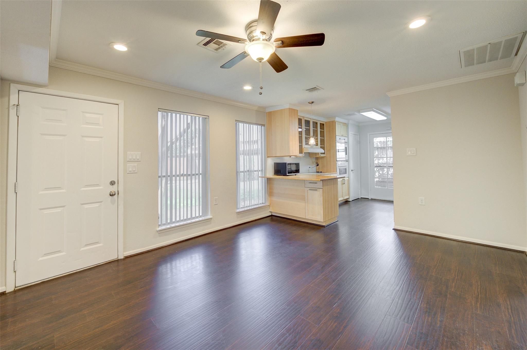 Empty living area with dark wood floors, vertical-blind windows, and an open kitchen with light wood cabinetry.