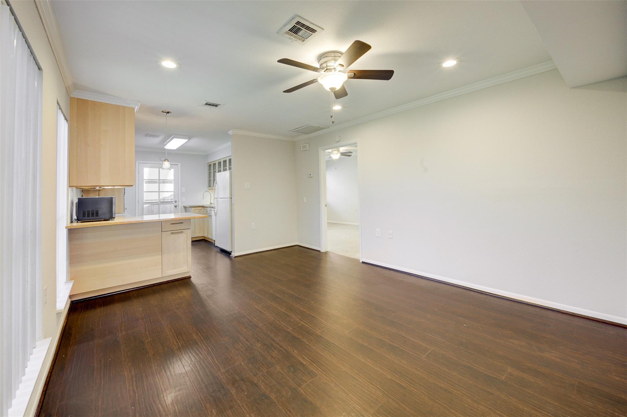 Empty living area with dark wood floors, ceiling fan, and an open kitchen with breakfast counter.