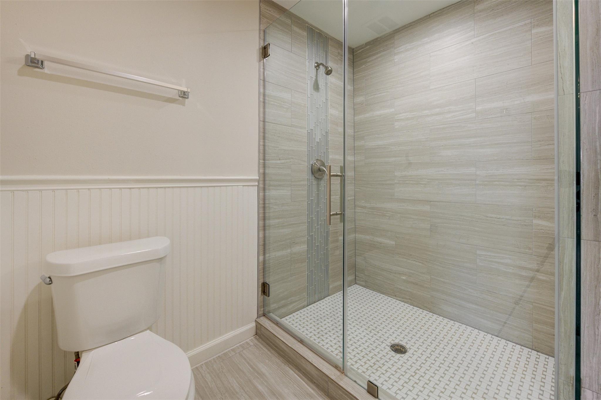 Bathroom with glass walk-in shower, mosaic tile floor, and toilet beside white wainscoting.
