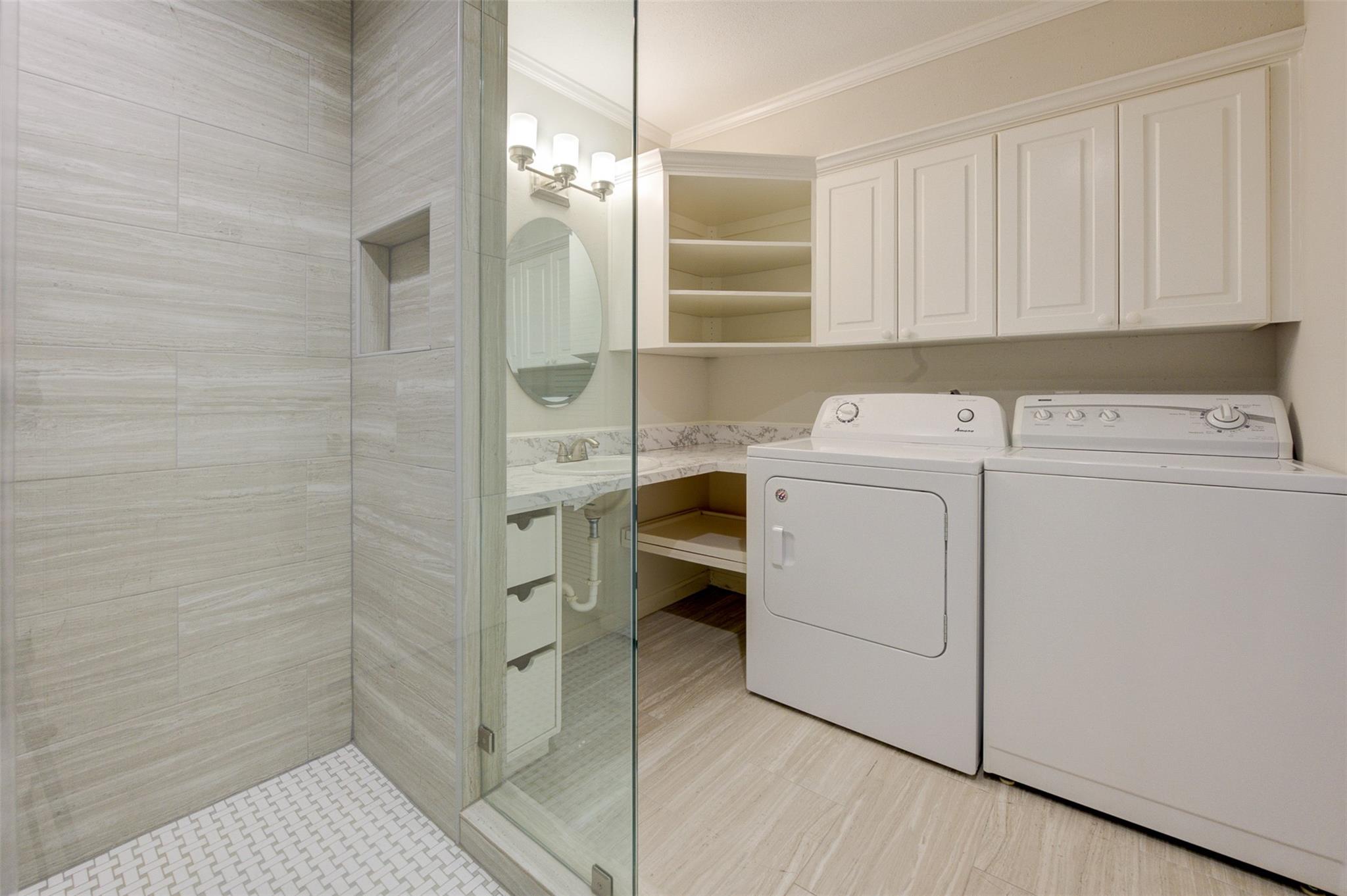 Laundry room with washer and dryer beside a glass-enclosed shower and white cabinetry.