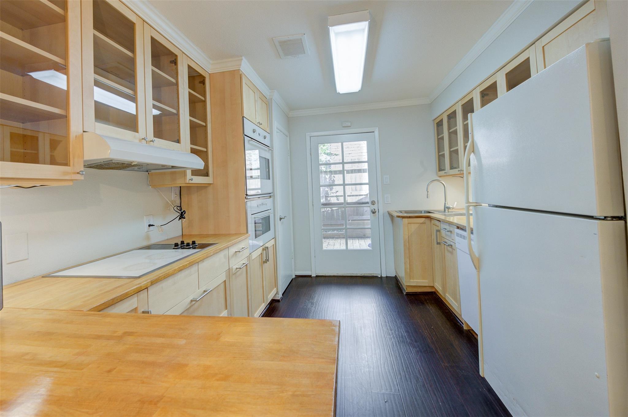 Galley kitchen with light wood cabinets, white appliances, dark flooring, and a glass-paneled door.