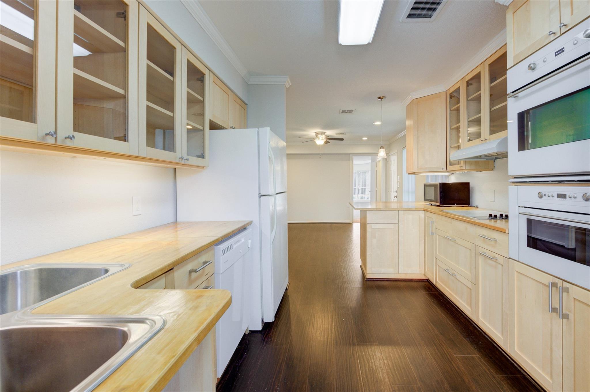 Galley kitchen with pale wood cabinets, glass-front uppers, double sink, white appliances, and dark flooring.