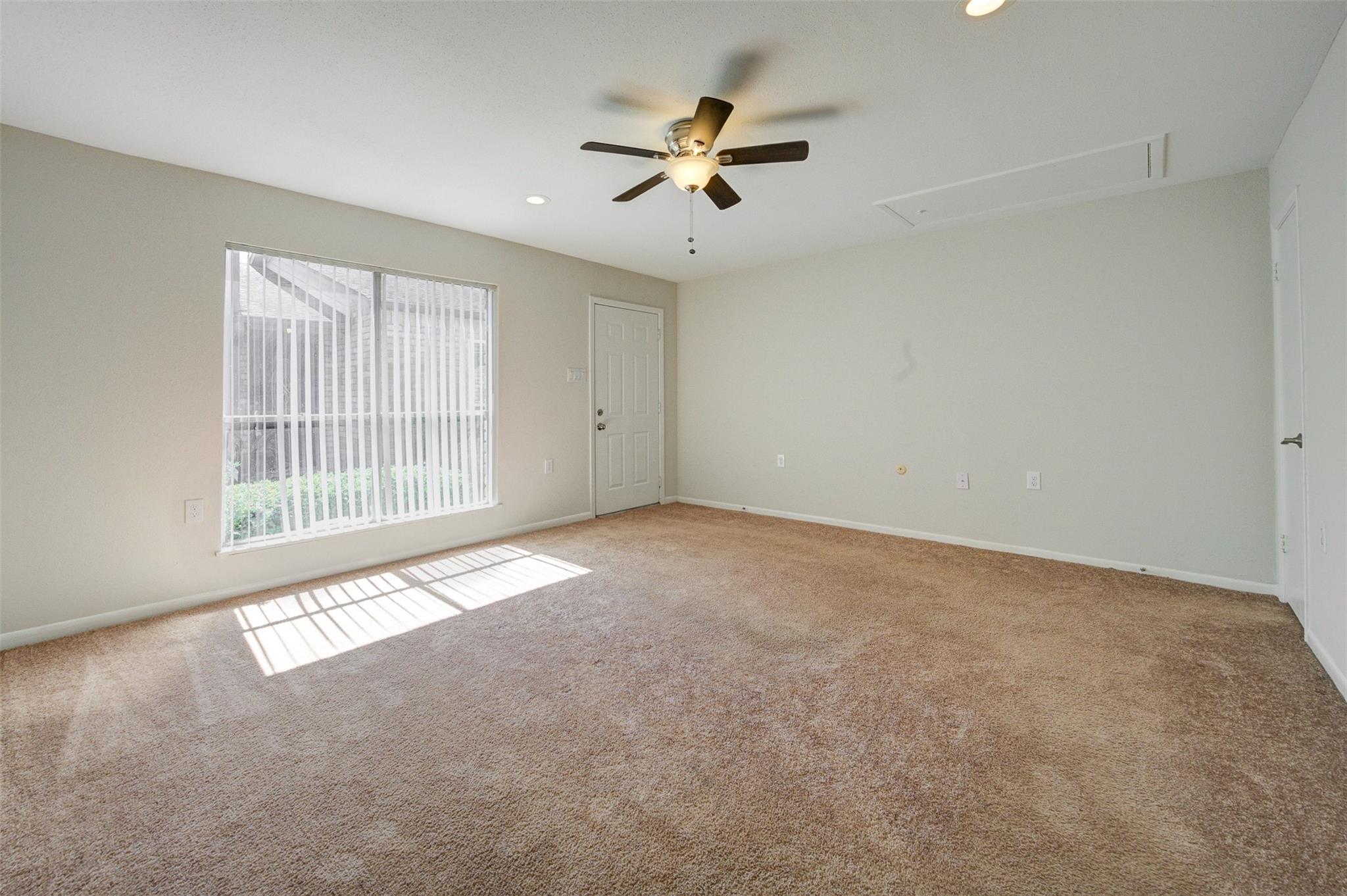 Empty carpeted bedroom with a ceiling fan and sunlight streaming through vertical blinds.