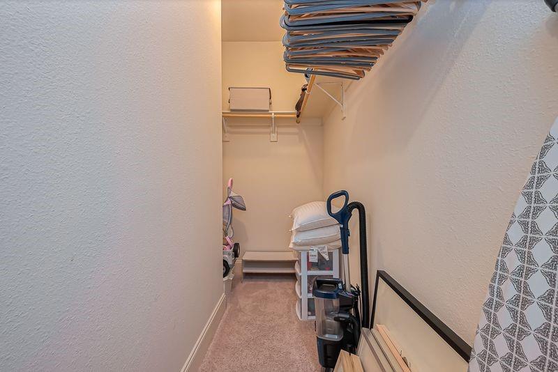 Walk-in closet with overhead clothes storage, shelving, stacked pillows, and a vacuum cleaner.
