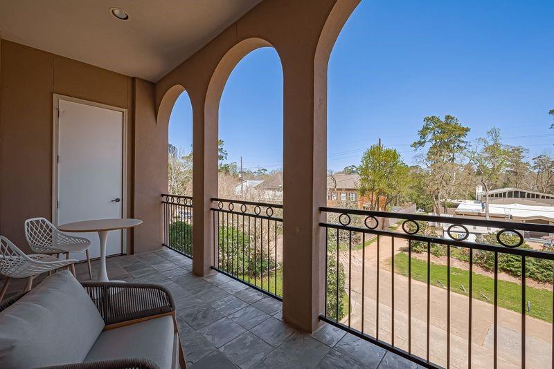 Covered apartment balcony with arched openings, tiled floor, and seating overlooking trees and neighboring buildings.