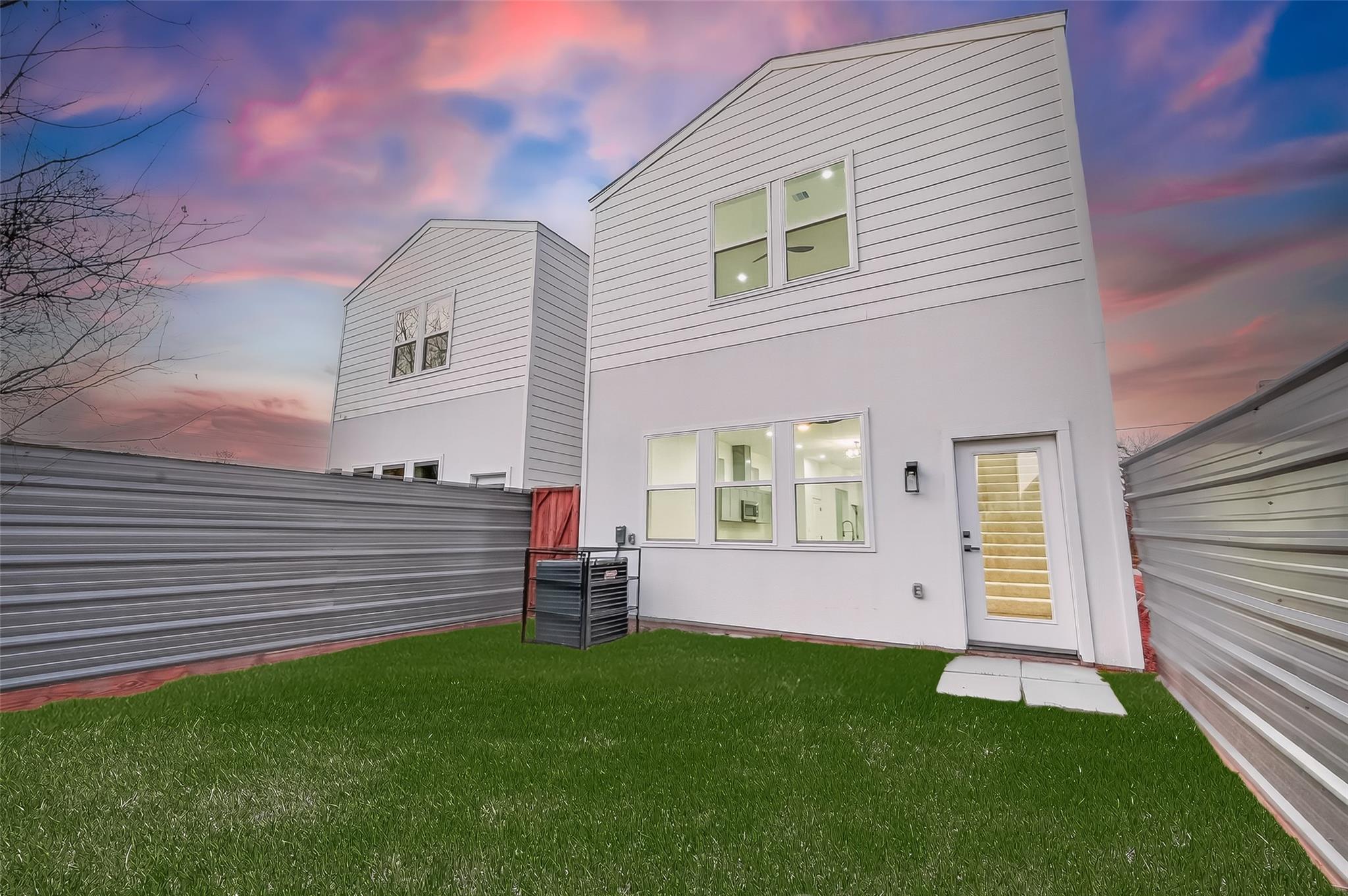 Modern two-story home with lit windows, a fenced lawn, and a glass-panel door at dusk.