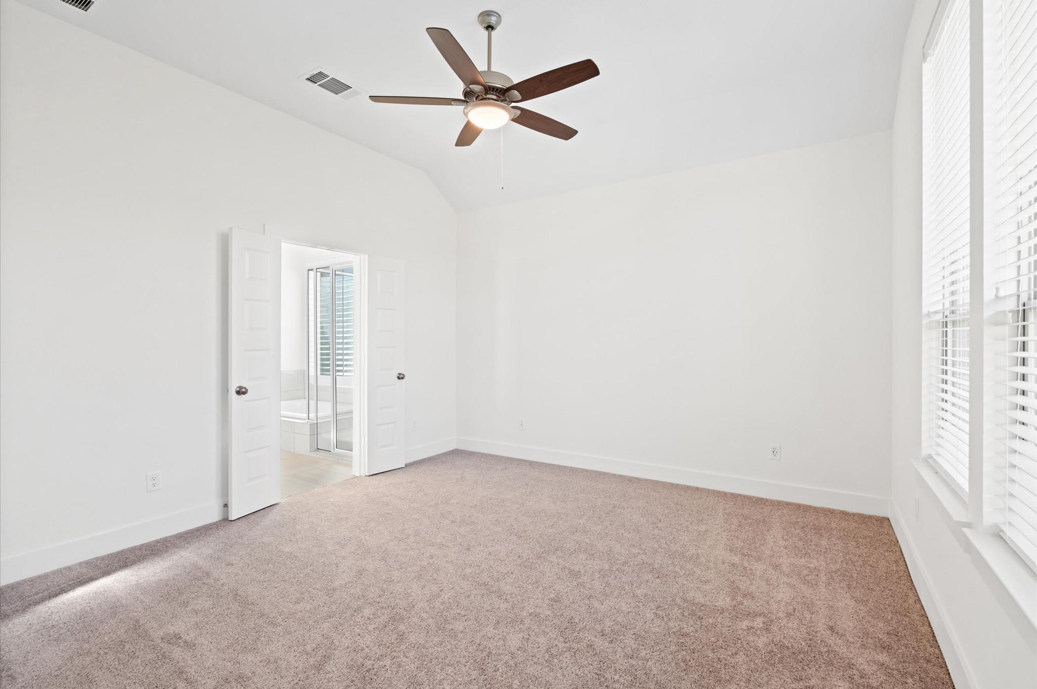 Empty carpeted bedroom with a ceiling fan, wide windows, and open doors to a bathroom.