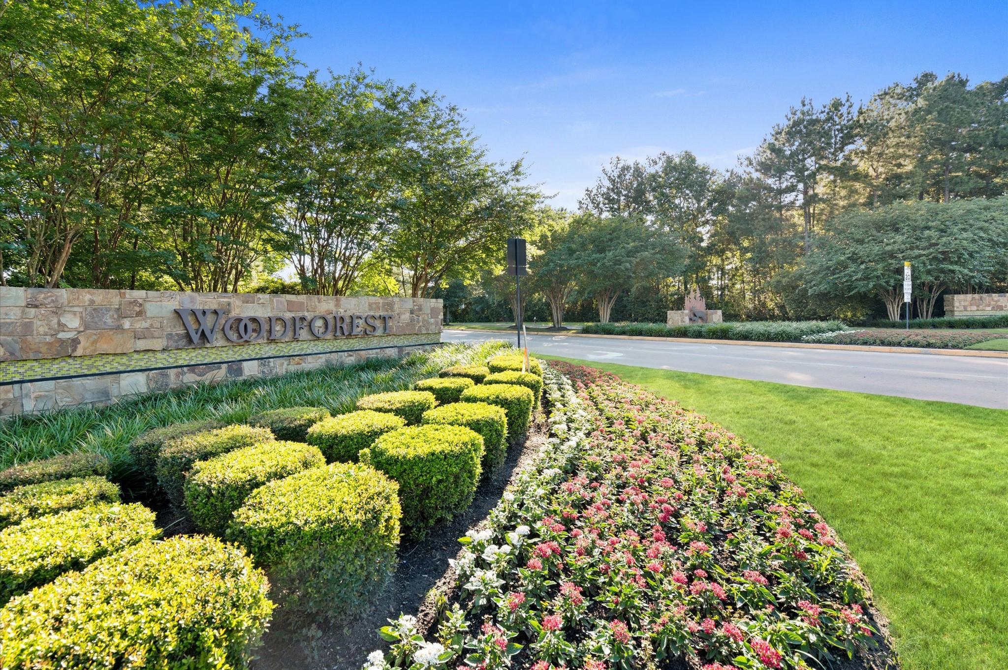 Woodforest community entrance sign beside landscaped flower beds and a tree-lined road.