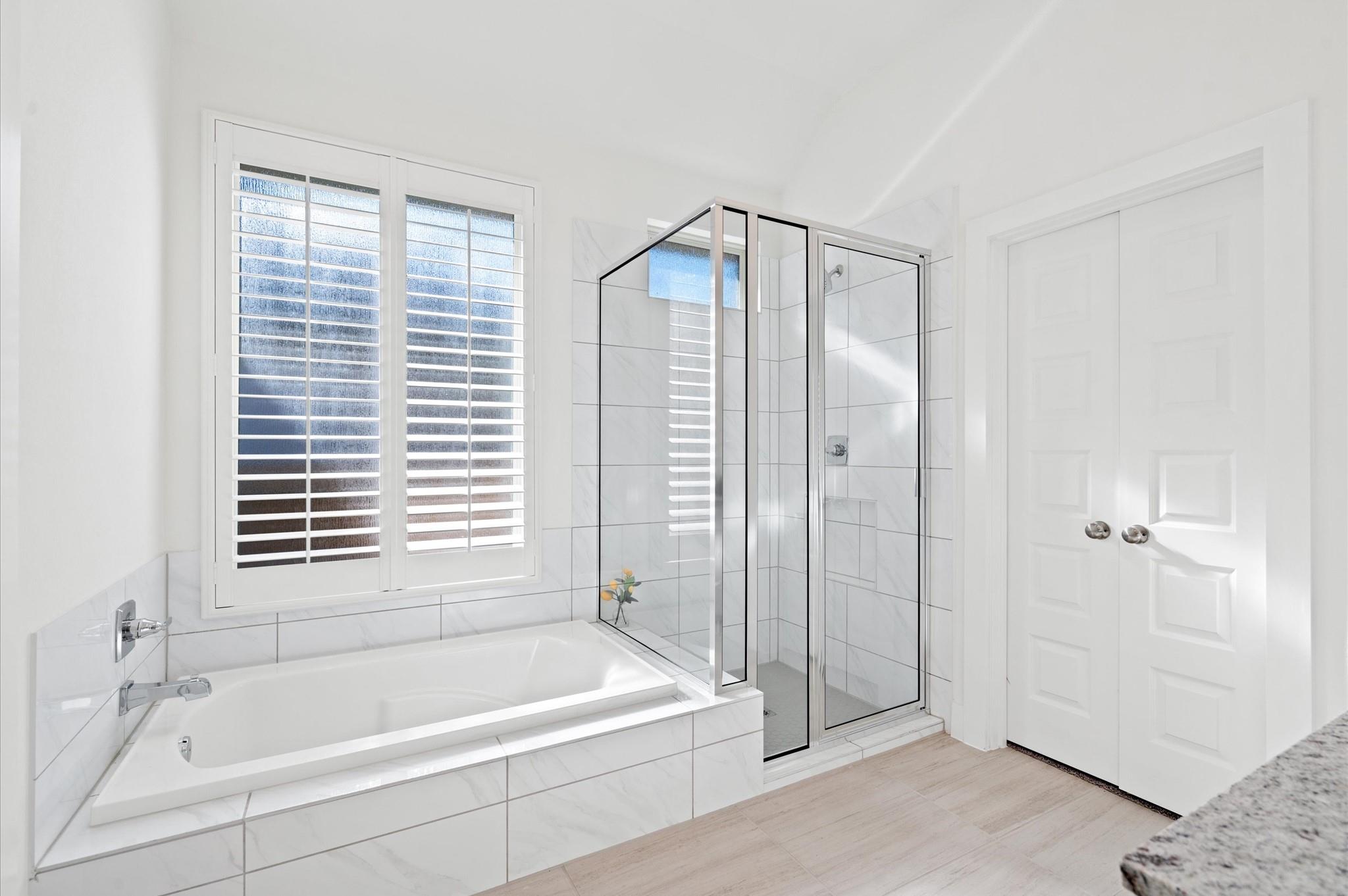 Bathroom with a white soaking tub, glass-enclosed shower, and plantation-shuttered window.