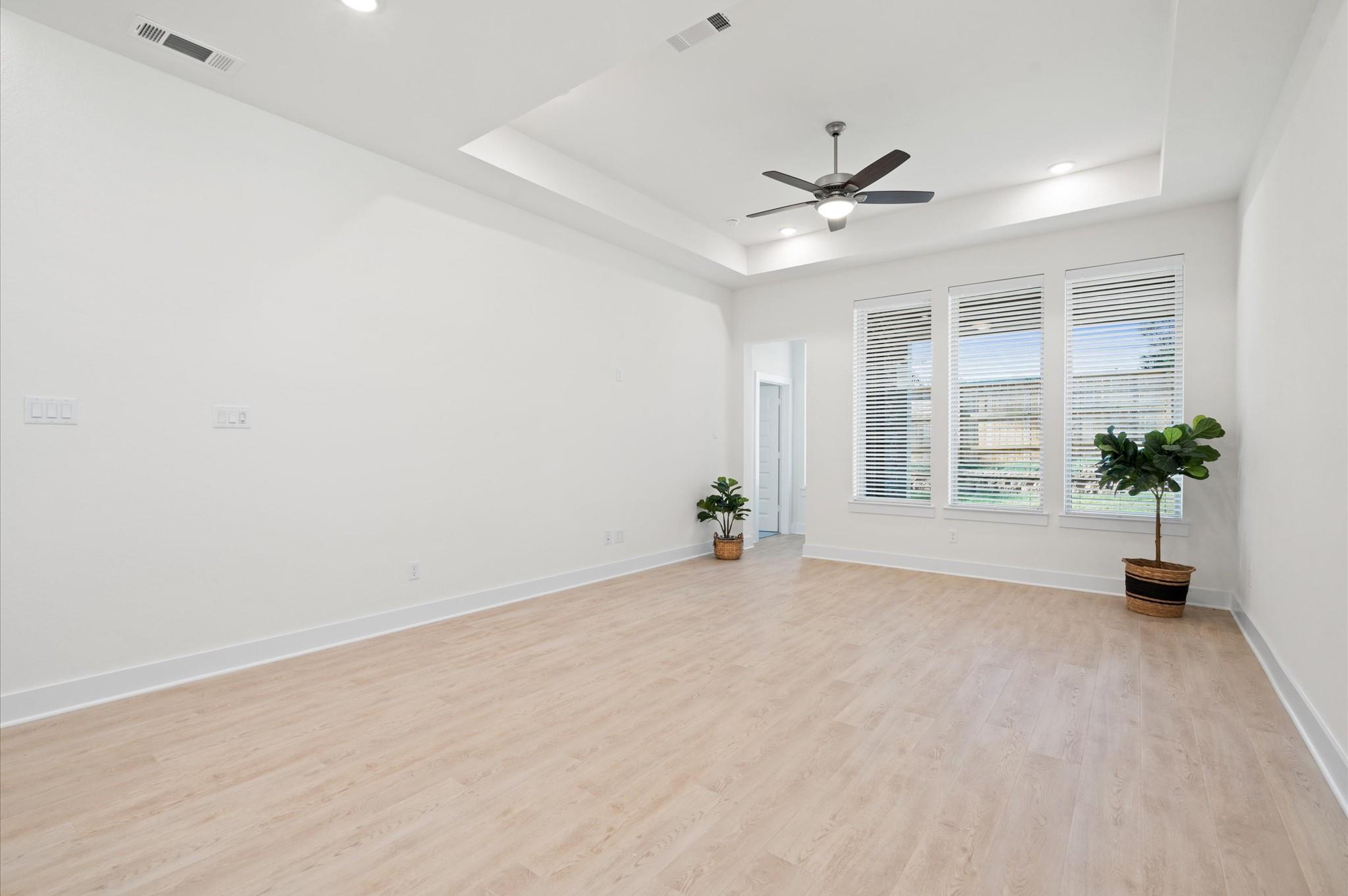 Vacant room with pale wood floors, a tray ceiling, ceiling fan, three tall windows, and potted plants.