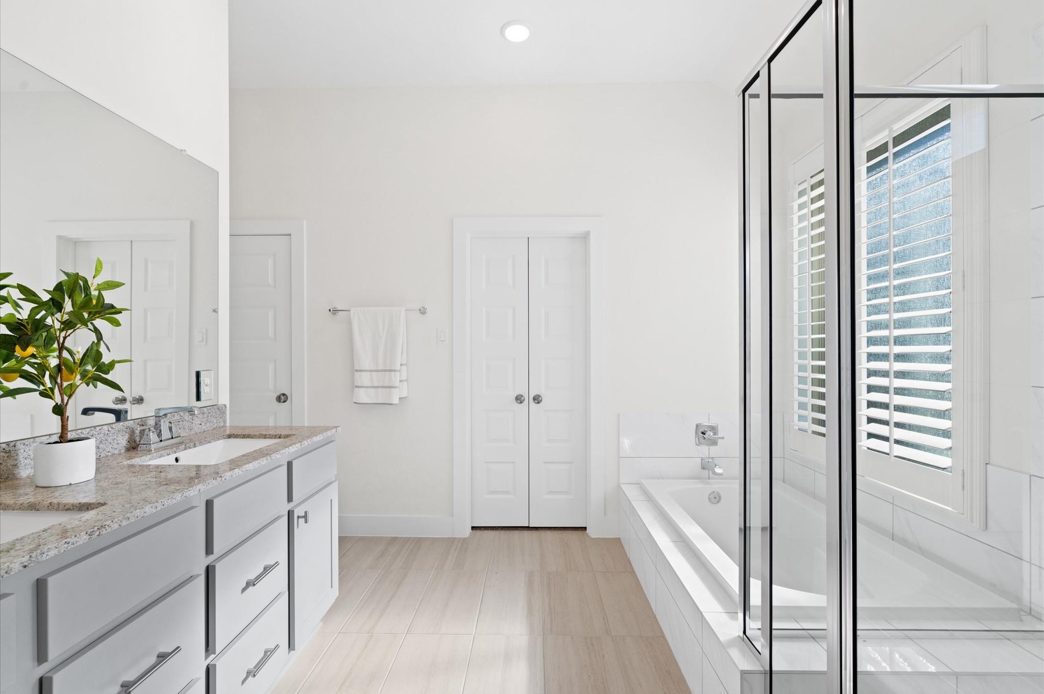 Bright bathroom with granite vanity, glass shower, bathtub, and shuttered window.