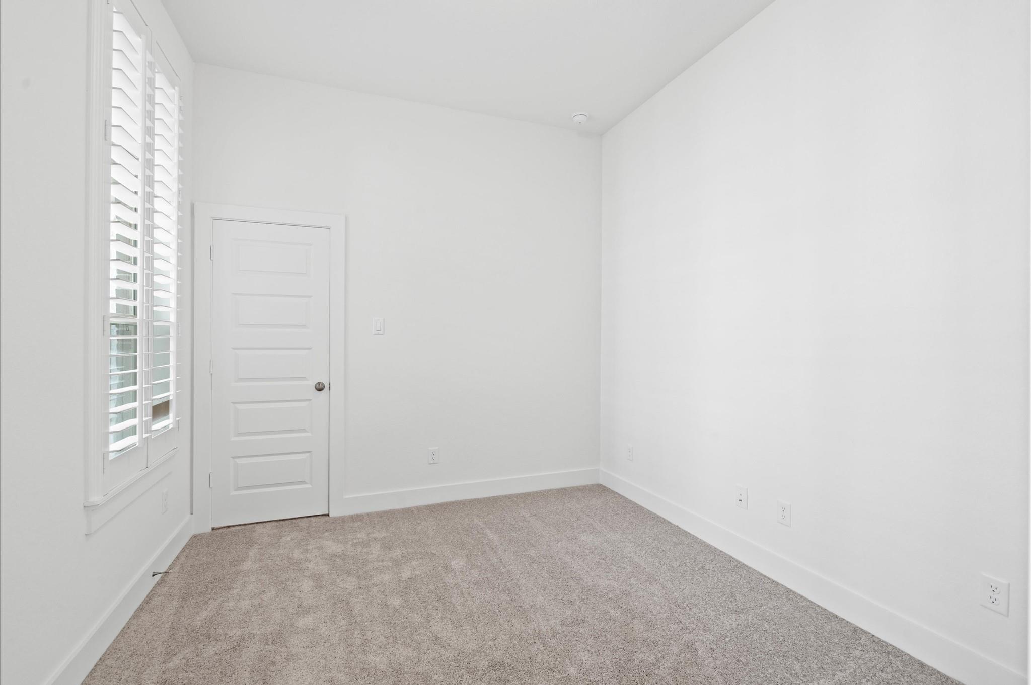 Empty carpeted room with white plantation shutters and a paneled door.