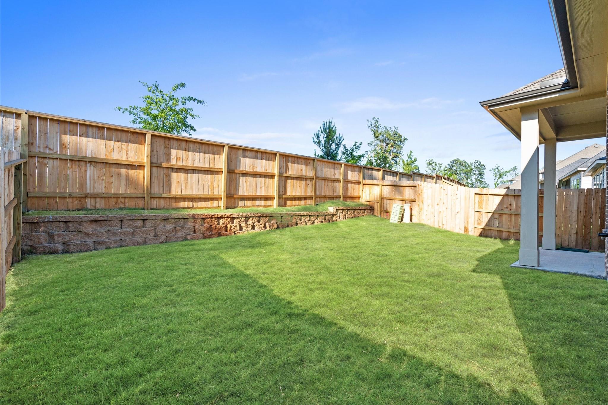 Fenced backyard with a broad lawn, tiered retaining wall, and covered patio.