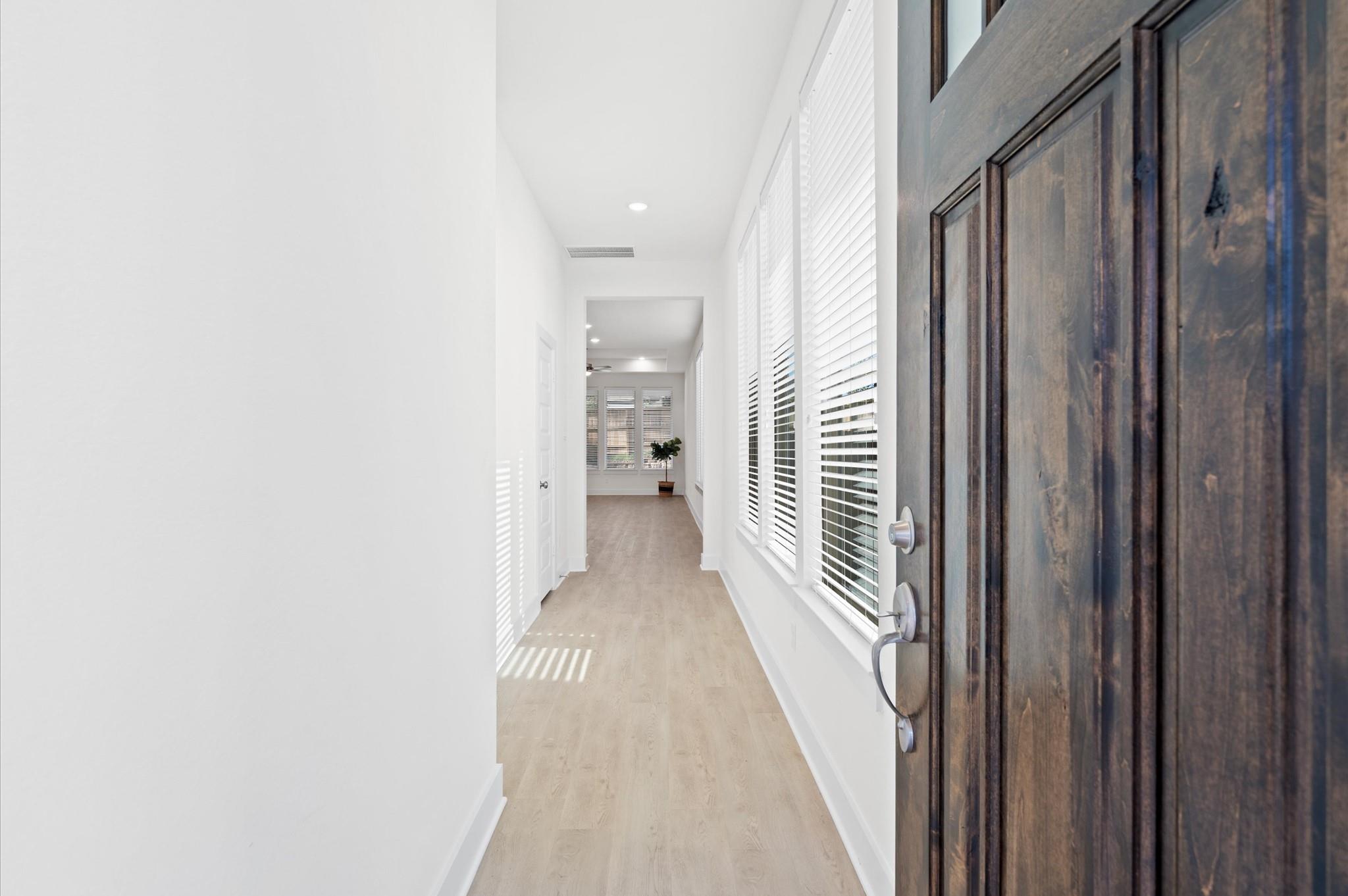 Entry hallway with light wood floors, tall blinded windows, and an open dark wood front door.