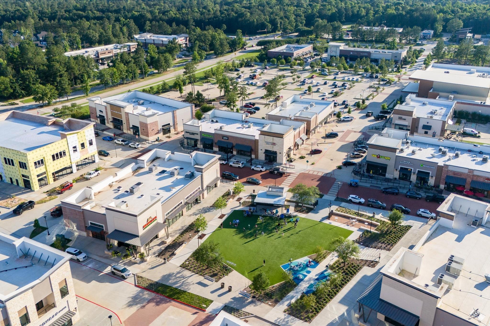 Aerial view of a shopping center with a central lawn, pavilion, and splash pad.