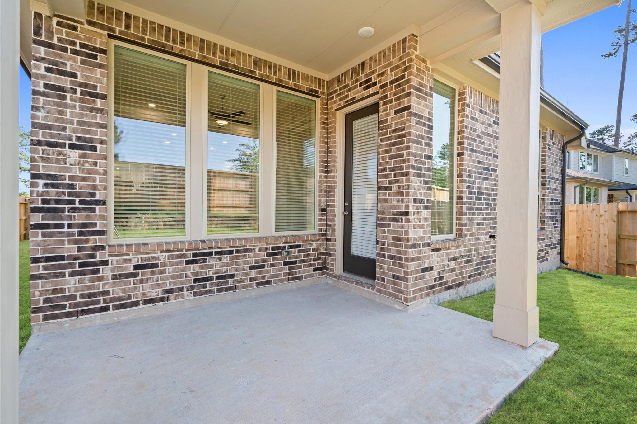 Covered concrete patio beside a brick home, with large windows and a fenced lawn.